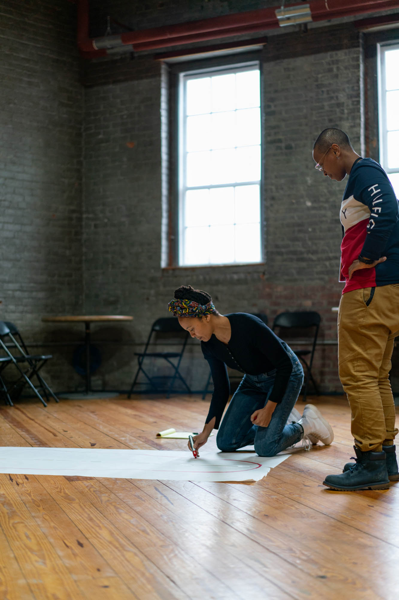 a black woman in jeans and a headwrap lays on white butcher paper as another black woman outlines her silhouette around her with a sharpie
