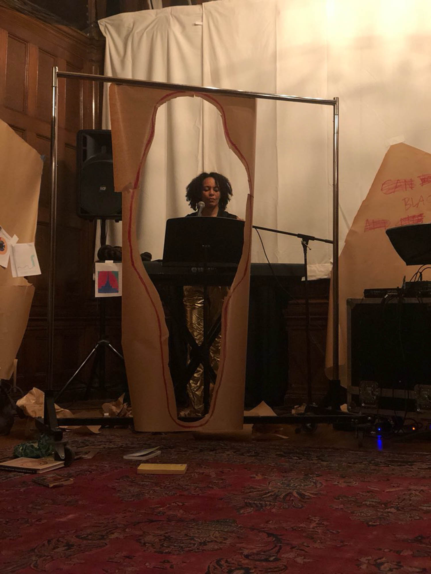 a black woman stands at a microphone and keyboard in gold metallic pants visible through a cut paper silhouette portal between captivity and freedom