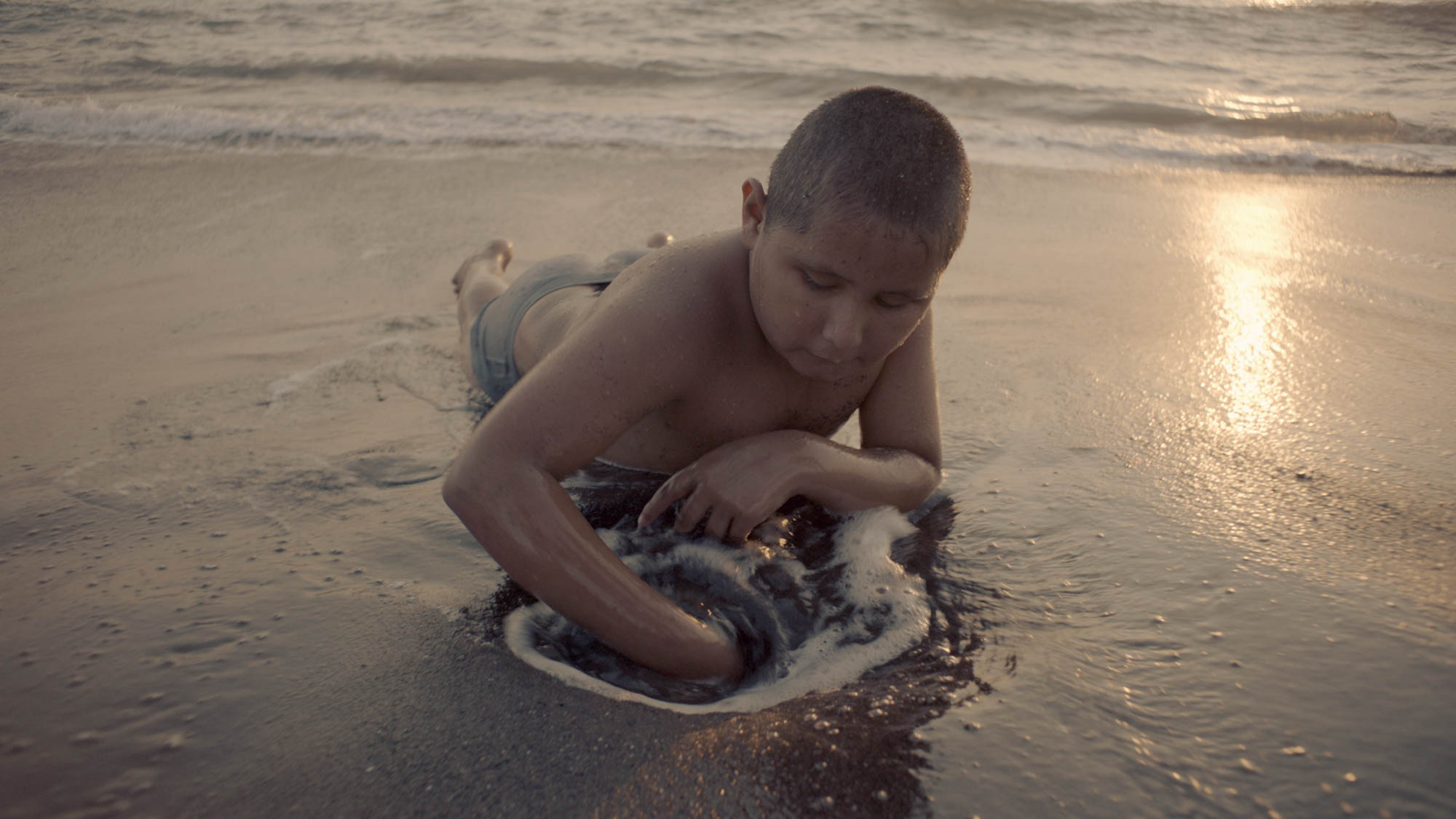 A young boy, about nine years-old is lying on a beach, his face towards camera. With his right hand, he digs a hole in the wet, foamy sand.