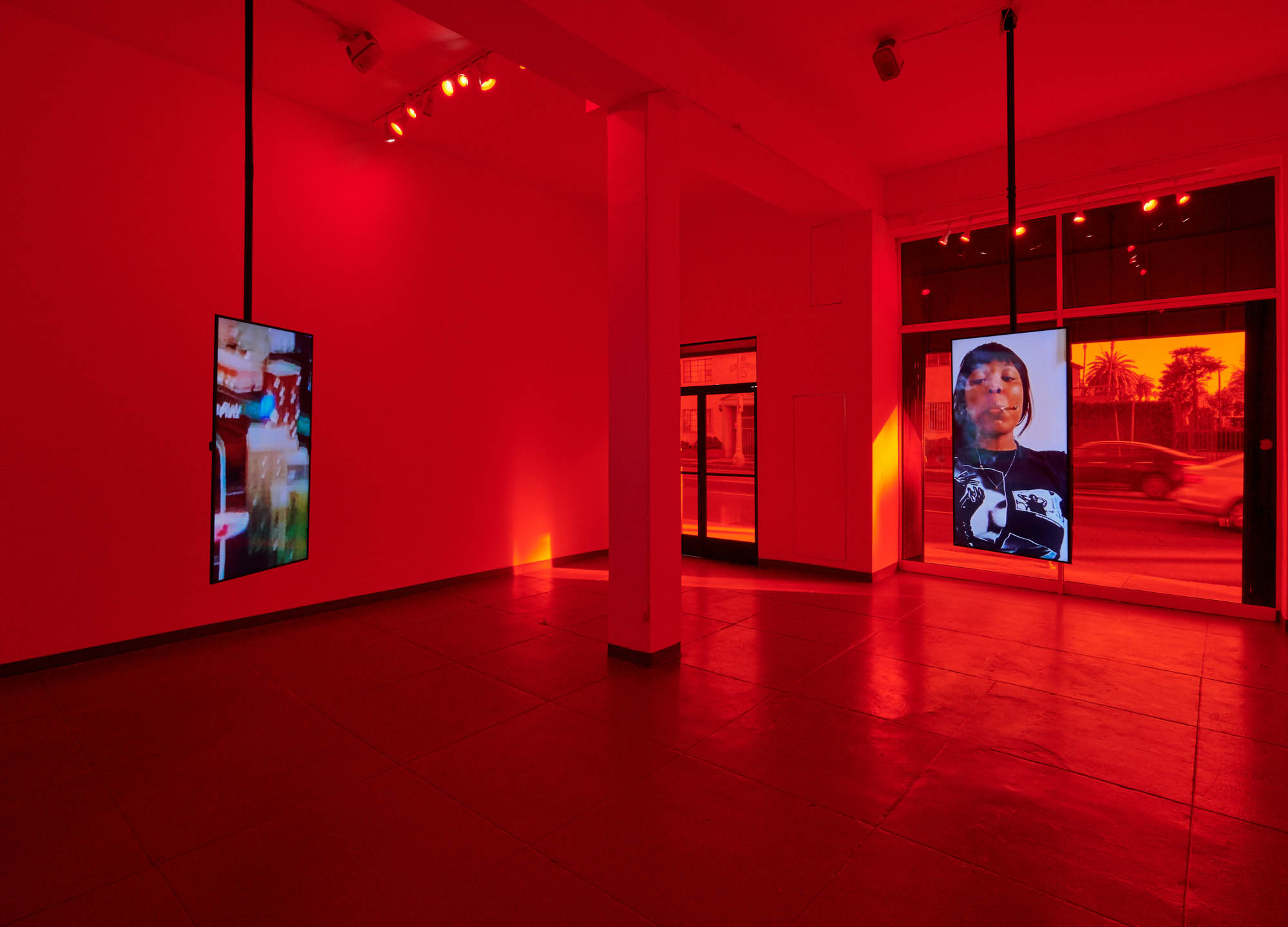 Large room with two monitors, one featuring the artist smoking a joint and the other featuring a blurry image, handgng from the ceiling in a red illuminated room.