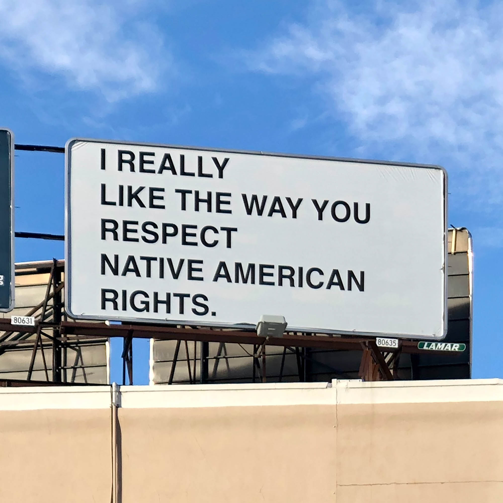 A large white billboard with black text sits atop a yellowish building with the brilliant blue sky and a few white clouds in the background.