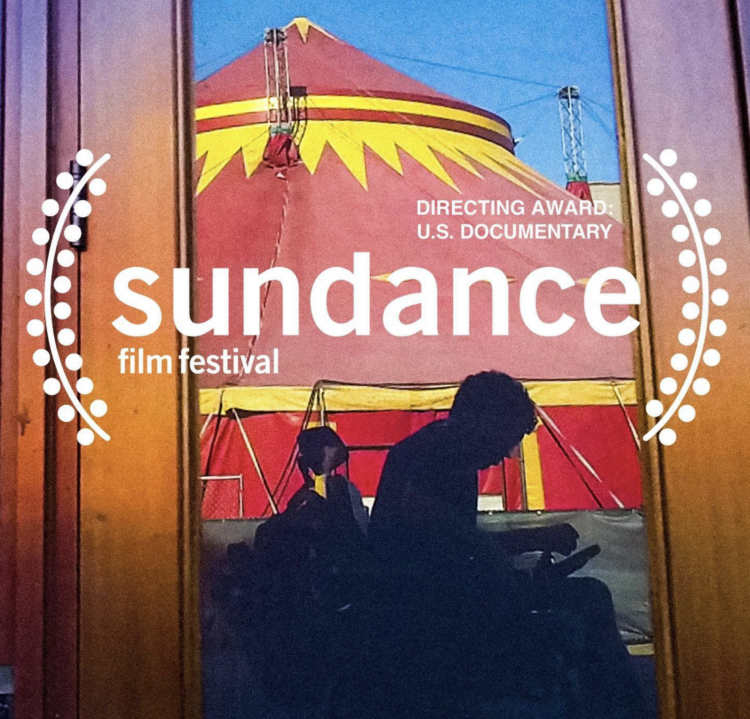 In a reflection of an unmarked storefront is a grayish silhouette of a man using an electric wheelchair. Overlaid on the photograph are white festival laurels that read “Directing Award: U.S. Documentary, Sundance Film Festival 2022