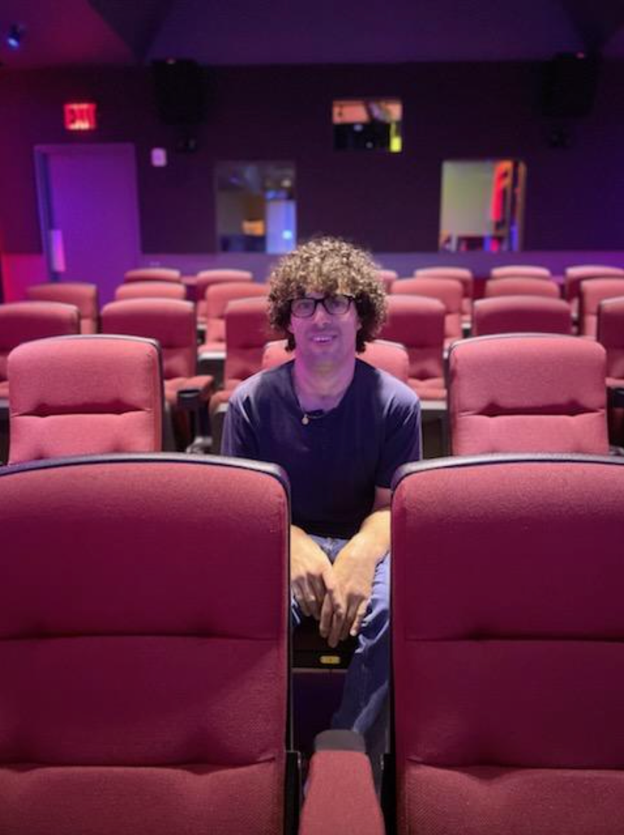 Reid, a white man with curly hair and glasses, sits in an empty, brightly lit theater and smiles subtly at the camera.