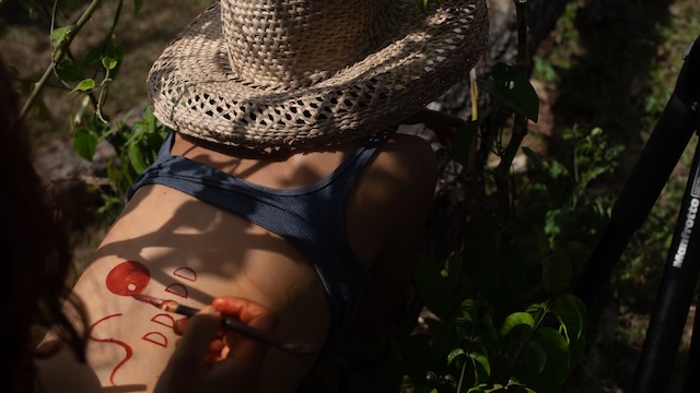 Someone paints the back of a woman wearing a wide-brim straw hat with red paint.