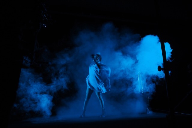 A performer on a blue, smoky stage looks to the side and covers their eyes with their hand.