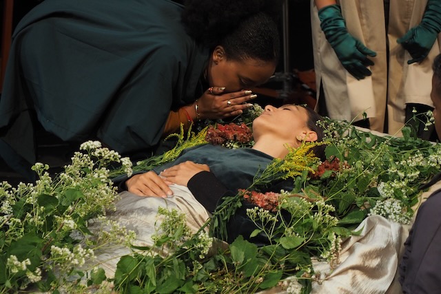 A woman whispers into the ear of another woman, lying down among flowers.