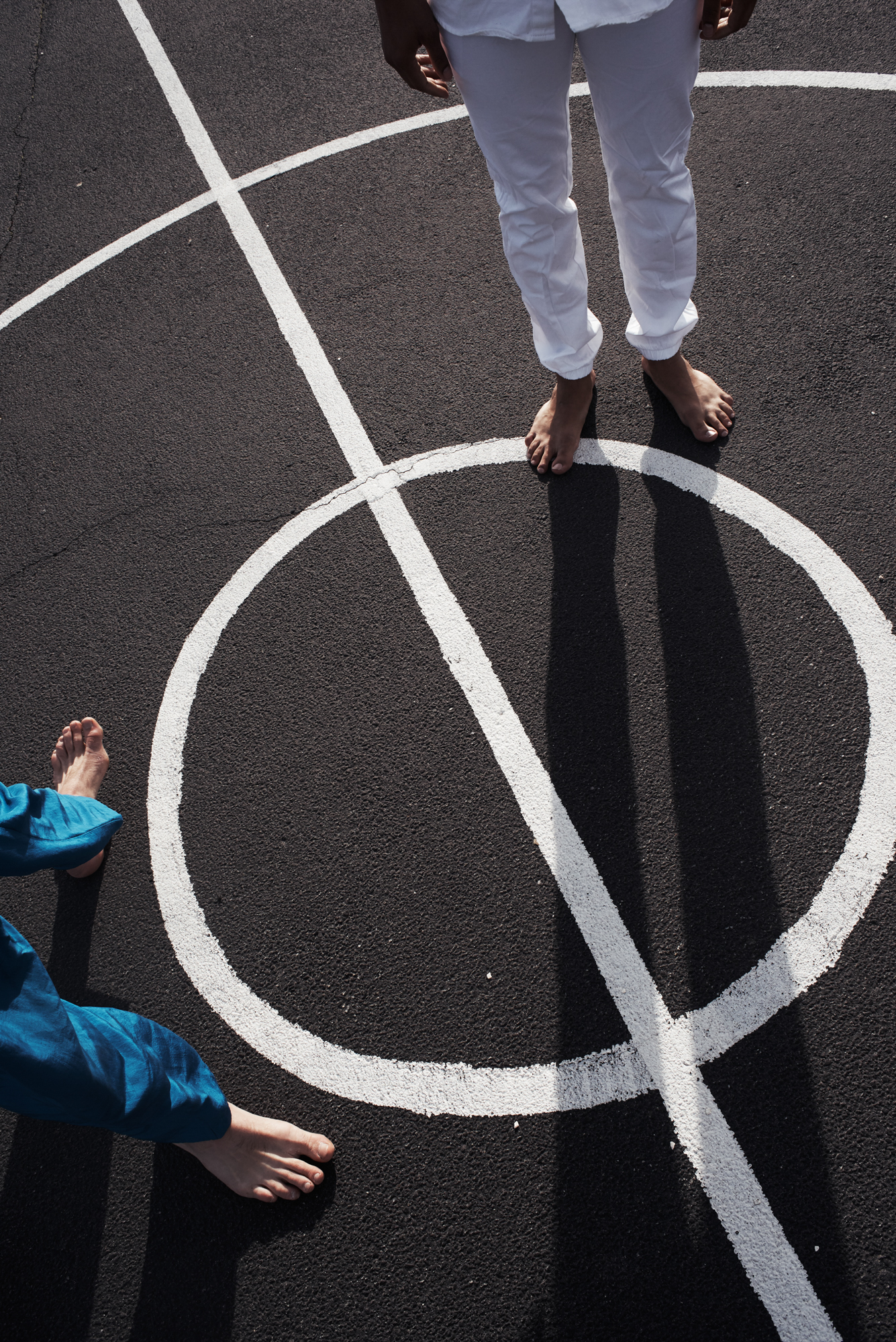 We see only the legs of 2 people who stand on black pavement painted with white concentric circles and a bisecting line. One person wears white and the other wears bright blue.