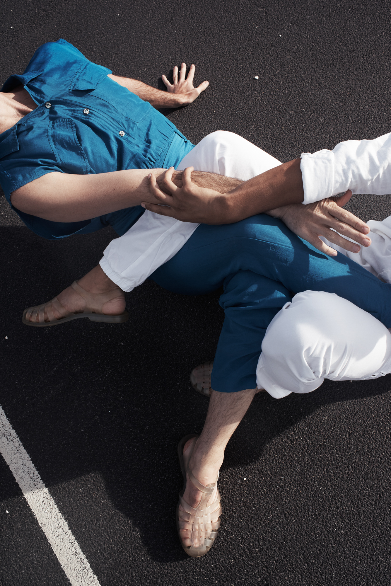 Two people, one wearing white, the other bright blue, sit on black pavement and wrap their legs around each other, arms clasped and arching away, with their faces out of frame.