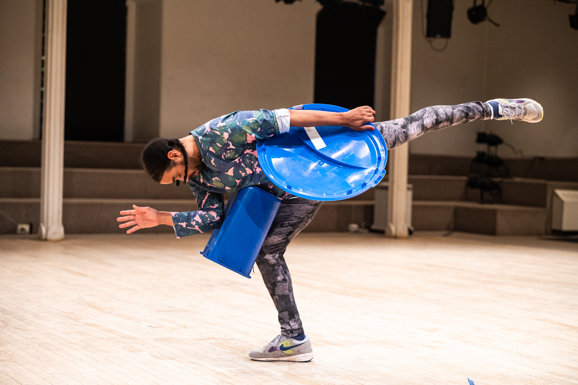 Rashaun, a Black person with dark hair pulled into 2 braids hovers over on one bent leg (arabesque), with a blue trash can sandwiched between his right elbow and right standing leg, and a blue trash lid held between his left arm and left hip, like a purse.