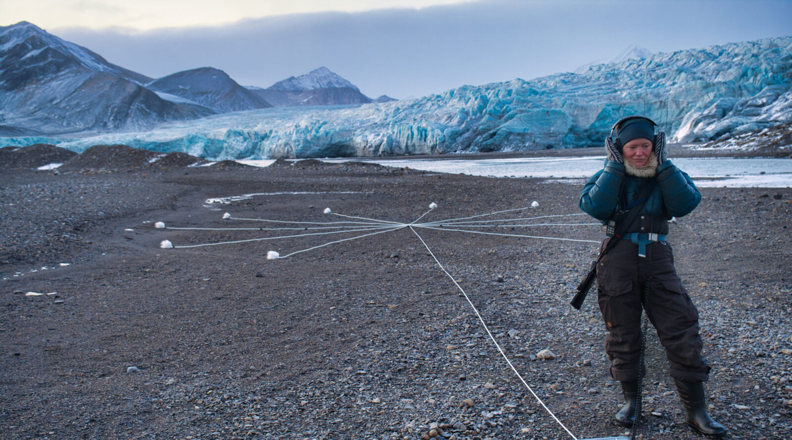 A barren landscape with sharp mountains and a large glacier behind an 11-spoked portable Macrophone; a woman in outdoor gear with a rifle is listening with headphones.