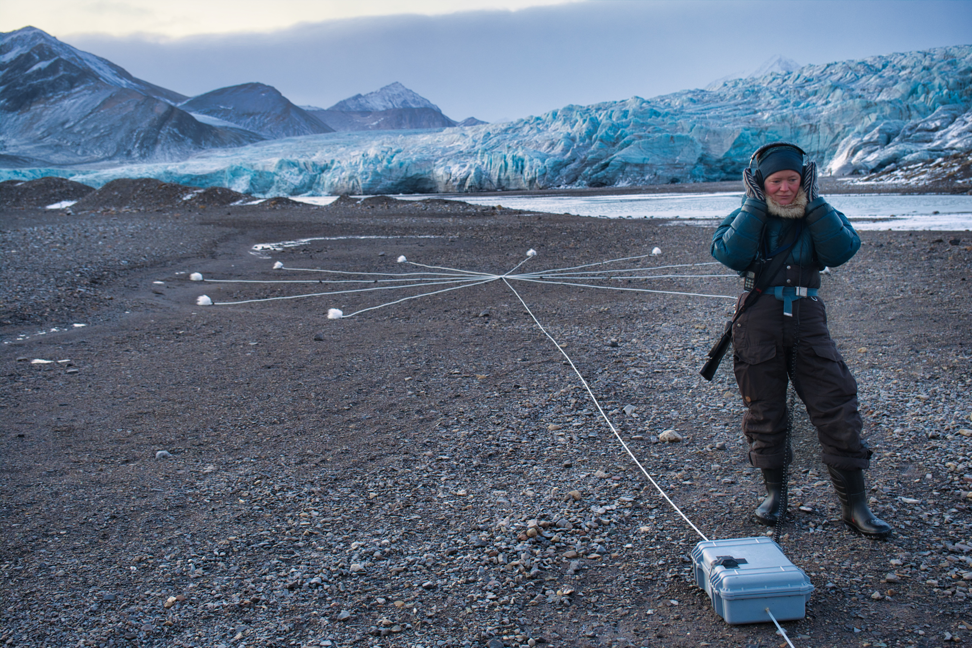 A barren landscape with sharp mountains and a large glacier behind an 11-spoked portable Macrophone; a woman in outdoor gear with a rifle is listening with headphones.