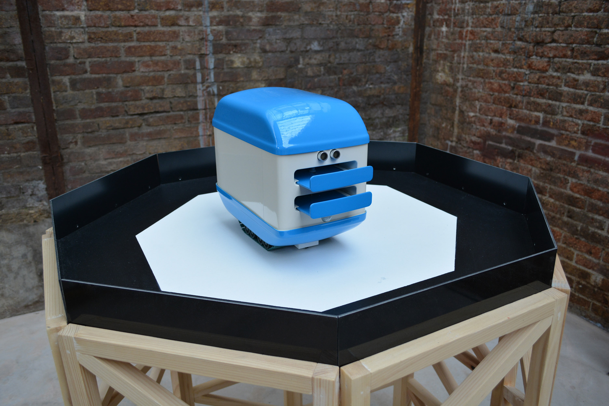 A photograph of a cute, bright blue, and off-white colored robot shaped like an electric toaster. The robot is sitting on top of an octagonal plinth made from unpainted wood. In the background is a rough brick wall. Two eye-like sensors protrude from the front of the robot above opening slots where slices of bread may be inserted, it gives the impression of a face. The robot has green plastic tank tracks to move around on.