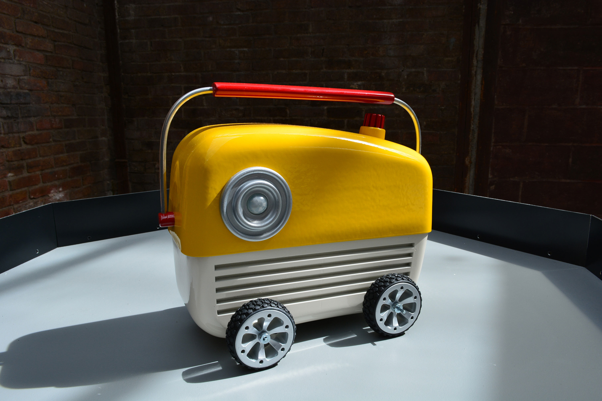 A photograph of a cute, bright yellow, and off-white colored robot shaped like a radio with a red plastic handle and a red dial. The robot is sitting on top of an octagonal plinth made from unpainted wood. In the background is a rough brick wall. An eye-like sensor protrudes from the front-left of the robot. Horizontal indentations across the front of the robot give the impression of a mouth. The robot has silver wheels with black tires to move around on.