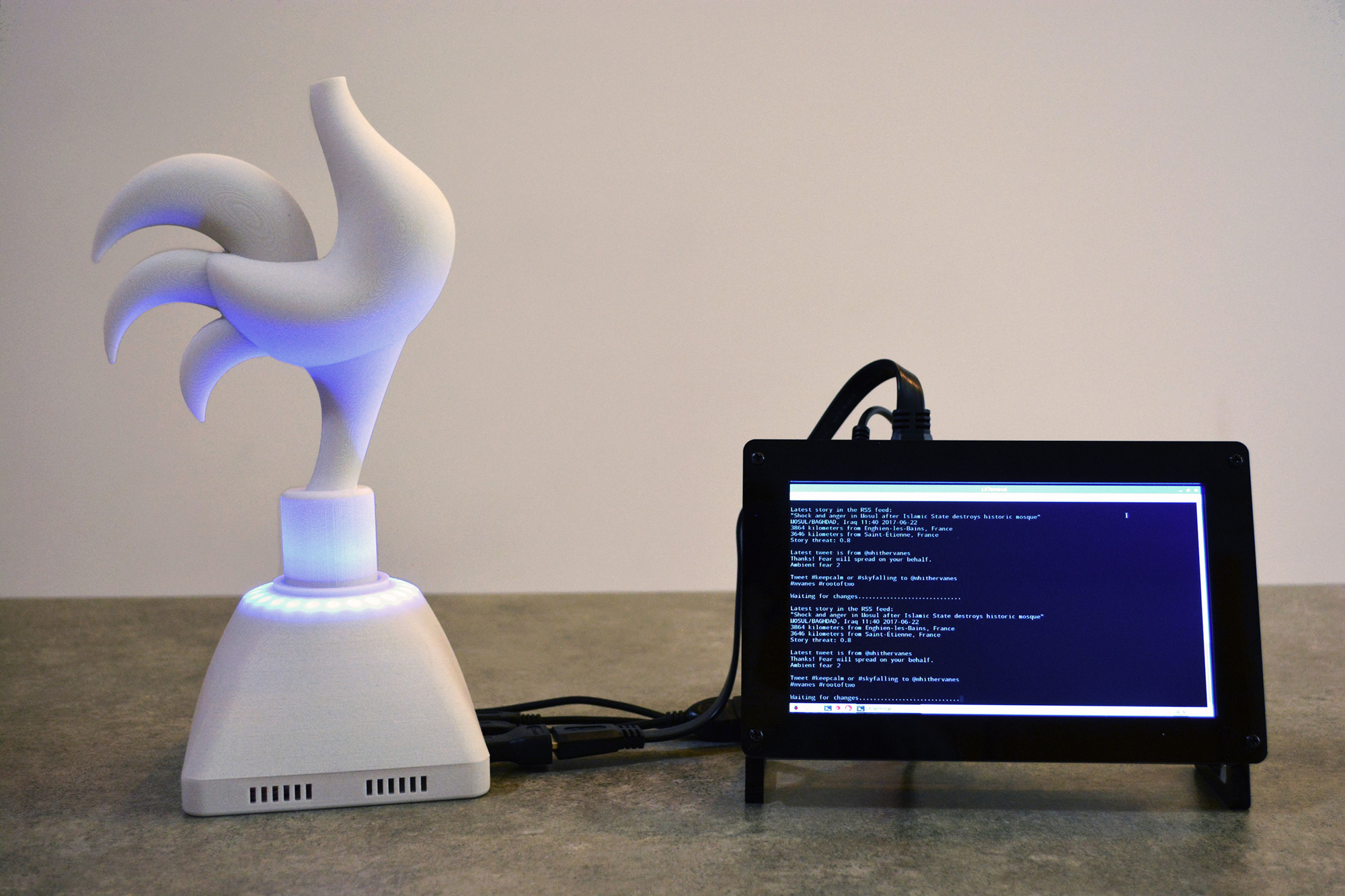 A photograph of a curvaceous, stylized sculpture of a rooster with no head mounted on top of a tapered base. It is against a white background and sits on a gray surface. The sculpture and the base are made of white plastic. Blue light is emitted from inside the base and uplights the sculpture with a blue glow. Cables run from the base and end in a black plastic framed monitor on which computer code is displayed.