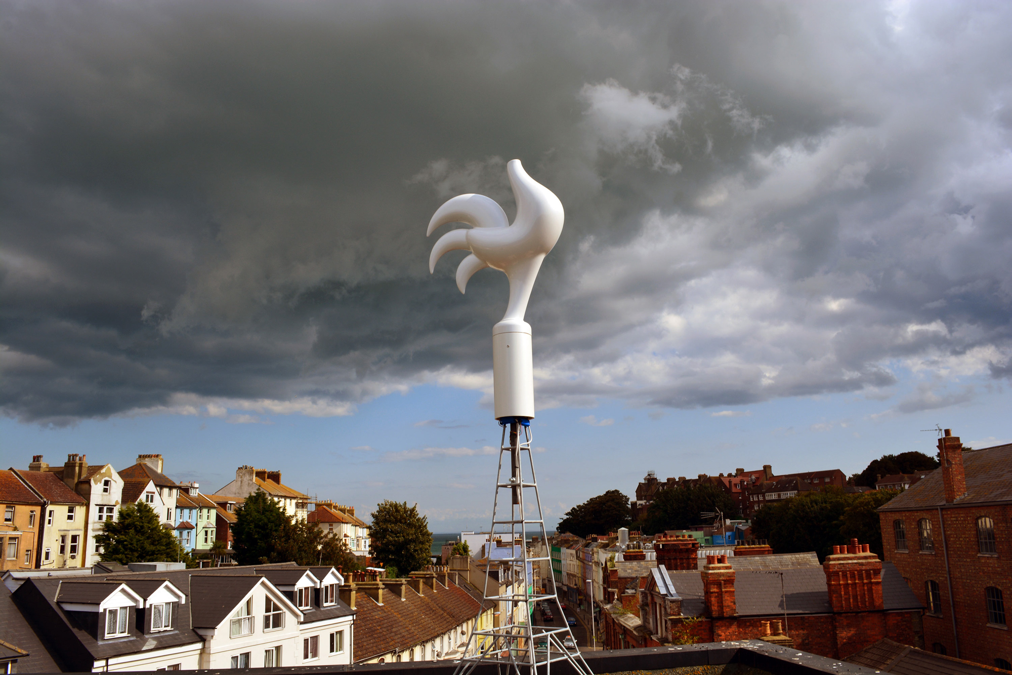 A photograph of a curvaceous, stylized sculpture of a rooster with no head mounted on top of a steel frame made in the proportions of the Eiffel Tower. In the background are the rooftops of an English seaside town. In the far distance on the horizon is the English Channel looking towards France. Overhead, gray storm clouds are forming in an otherwise blue sky.