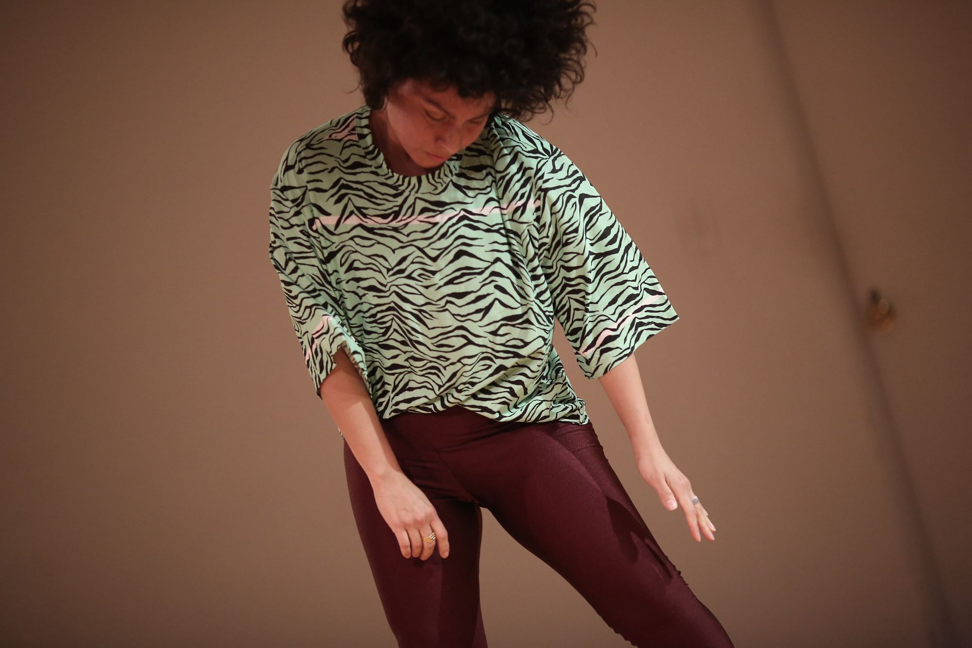 A Latinx woman with black curly hair and tan skin dances in a mint green and black tiger print t-shirt and maroon leggings. The floor is blonde wood, the walls are off-white her feet are bare.