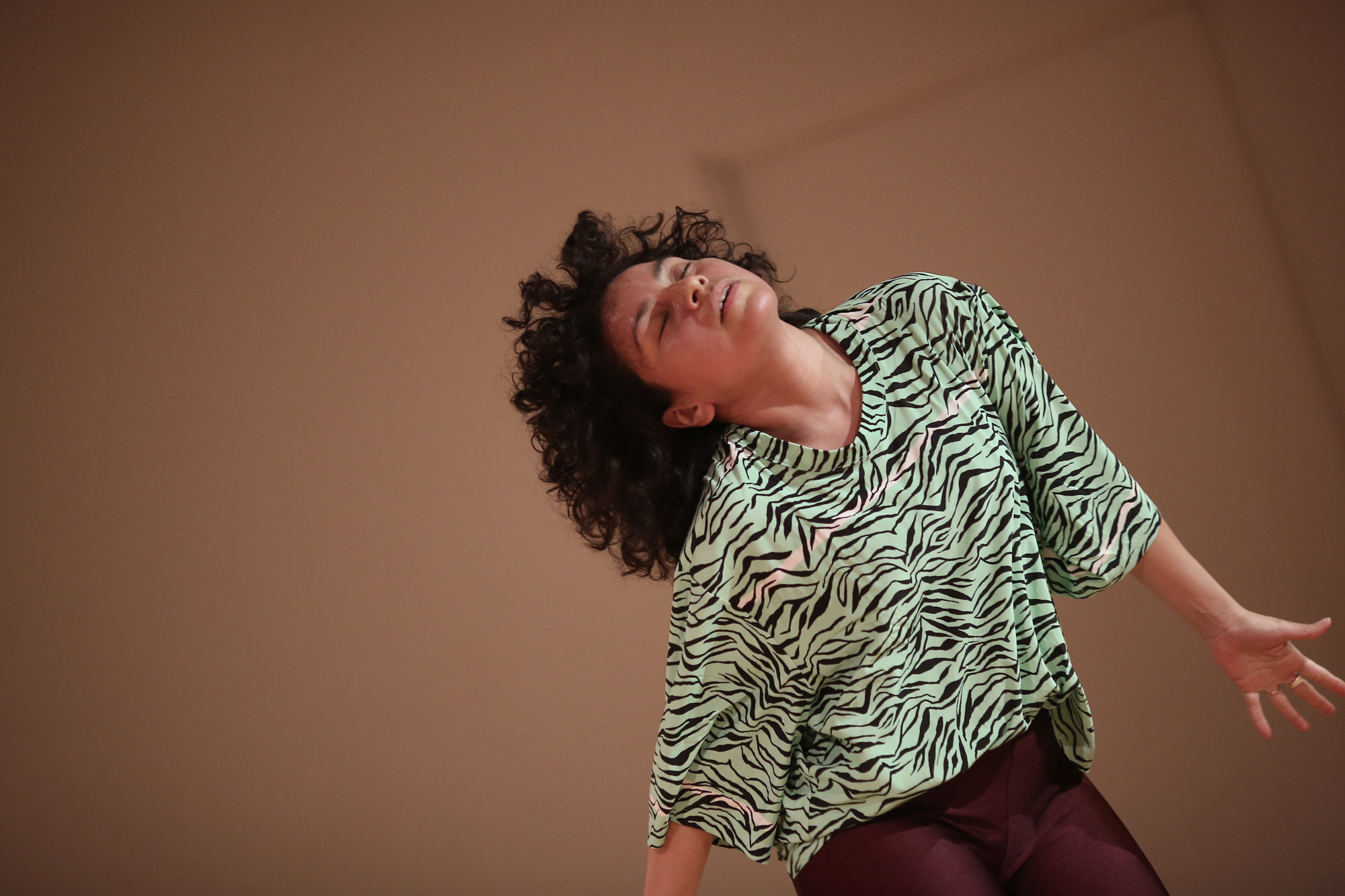 A Latinx woman with black curly hair and tan skin dances in a mint green and black tiger print t-shirt and maroon leggings. The floor is blonde wood, the walls are off-white her feet are bare.