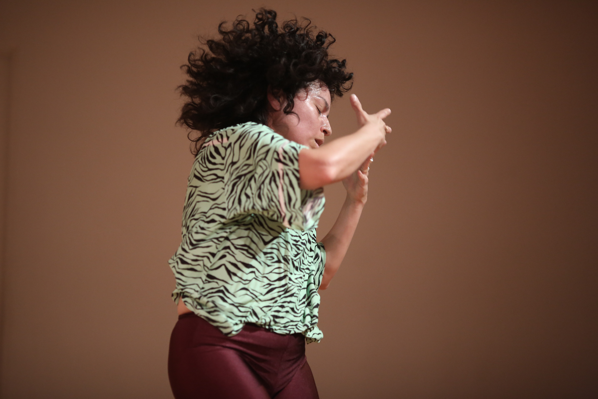 A Latinx woman with black curly hair and tan skin dances in a mint green and black tiger print t-shirt and maroon leggings. The floor is blonde wood, the walls are off-white her feet are bare.