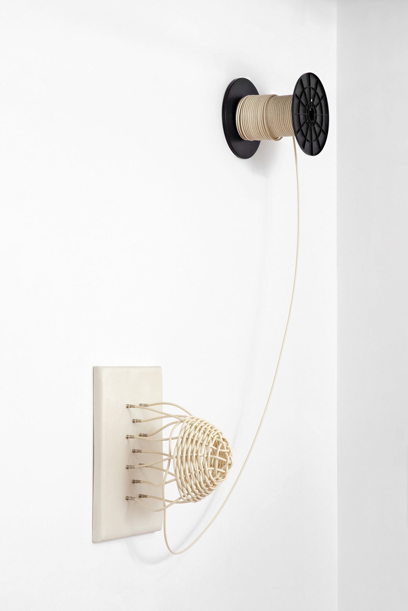 An oversized off-white rectangular ceramic utility wall plate sprouts a partly-woven basket joined to the plate through coaxial cable fittings; the same off-white coaxial cable that makes up the basket continues onto a large spool hung high on the wall.