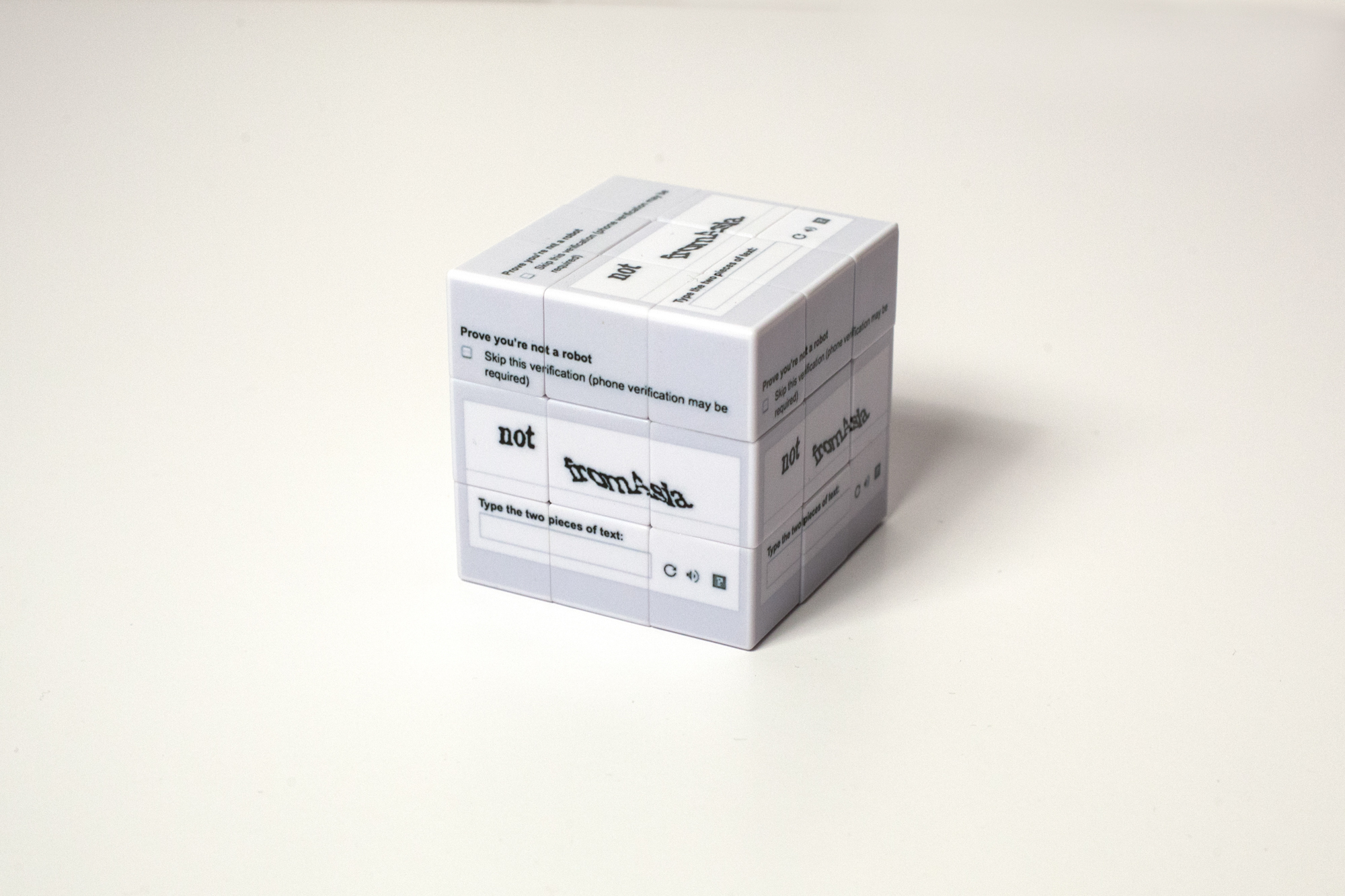 Shown on a white background, a white and grey Rubik’s cube is printed on all sides with a reCAPTCHA screenshot that instructs users to “Prove you’re not a robot” by entering the phrase “Not from Asia.”