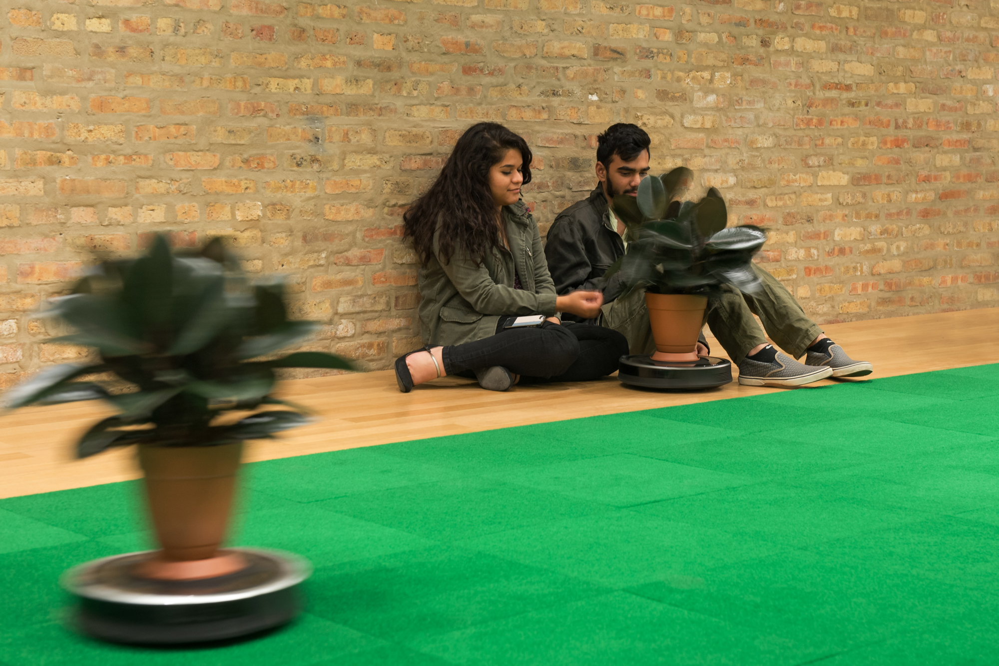 A young couple sits on the floor, leaning against an exposed brick wall, gently touching a potted rubber tree plant mounted on a Roomba vacuum that is brushing their knees, while another Roomba-mounted plant whirs over bright green carpet cutting diagonally through the foreground of the frame.