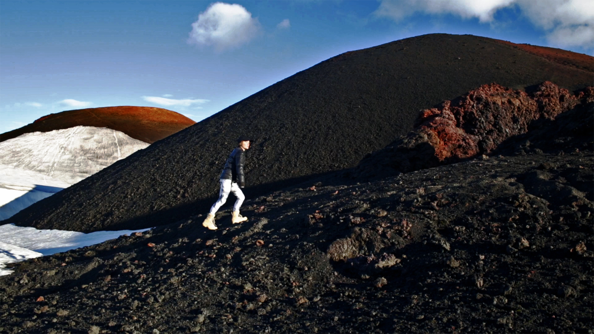4. Deep black, volcanic mountain peaks rise against a bright blue sky, here and there covered by snow and bright red pumice. Between them climbs a small, white person wearing black leggings, heavy boots and a black jacket. Their face turns at an angle towards the camera, looking off into the distance.