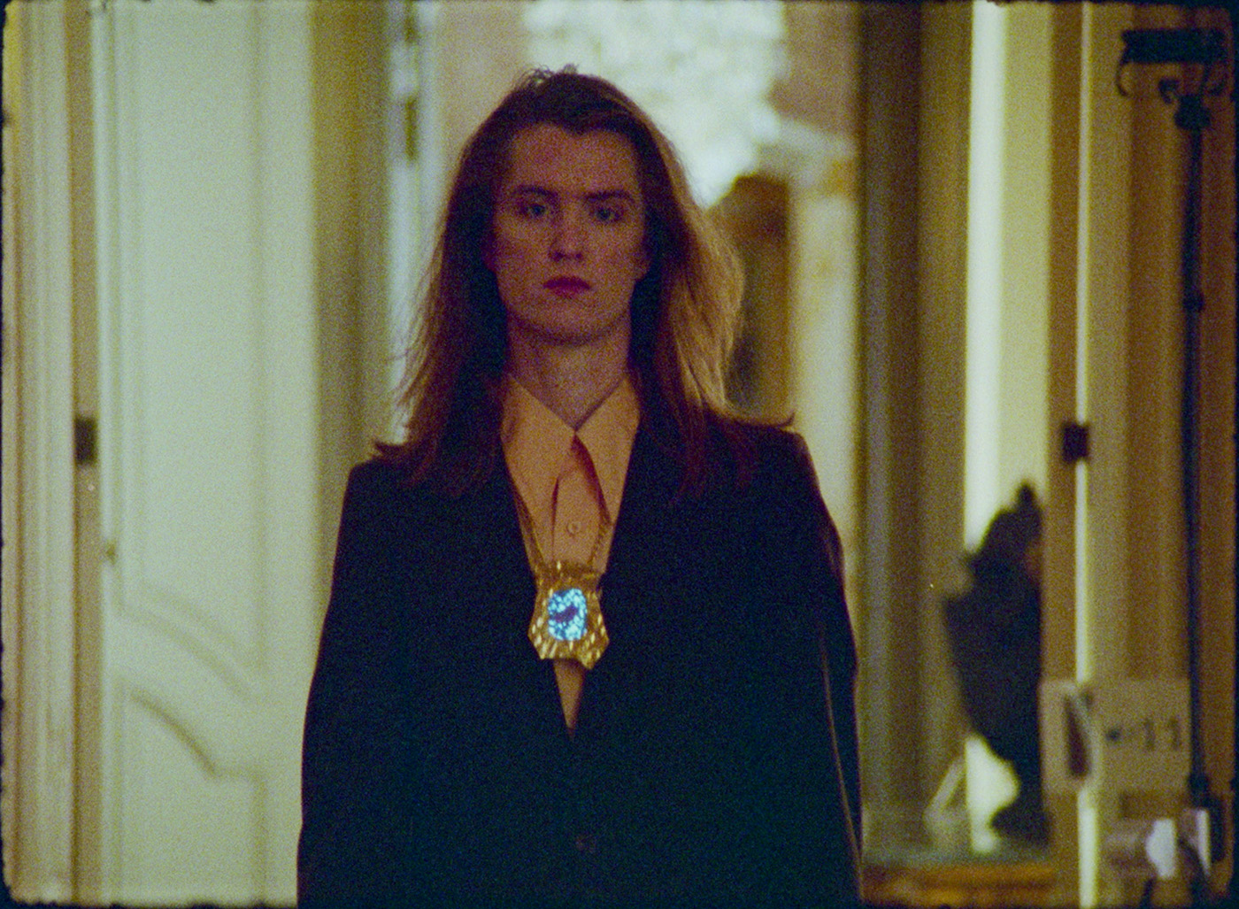 6. A slender, gender queer person with long, flowing blonde hair strides towards the camera, visible from waist up. They emerge from a grandiose architecture with a crystal chandelier blurred in the background. They wear a yellow shirt with oversized collar, a velvet suit jacket, and a large, jagged brass amulet shining light. They gaze directly at the camera with cool confidence.