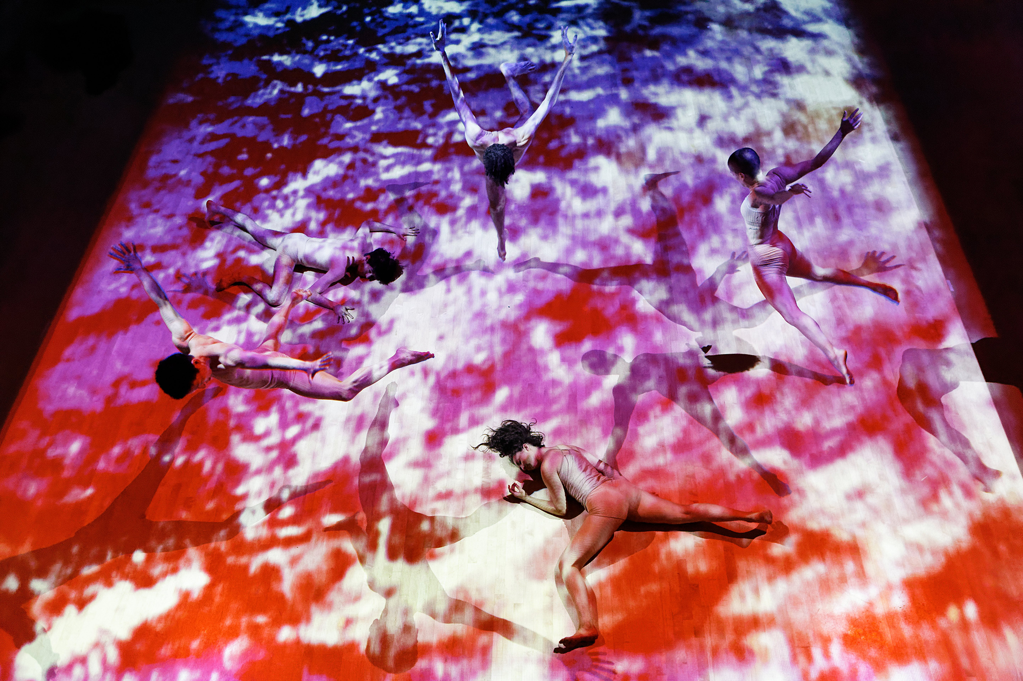Three performers jump, and two performers are on the floor under the red and purple abstract video projection.