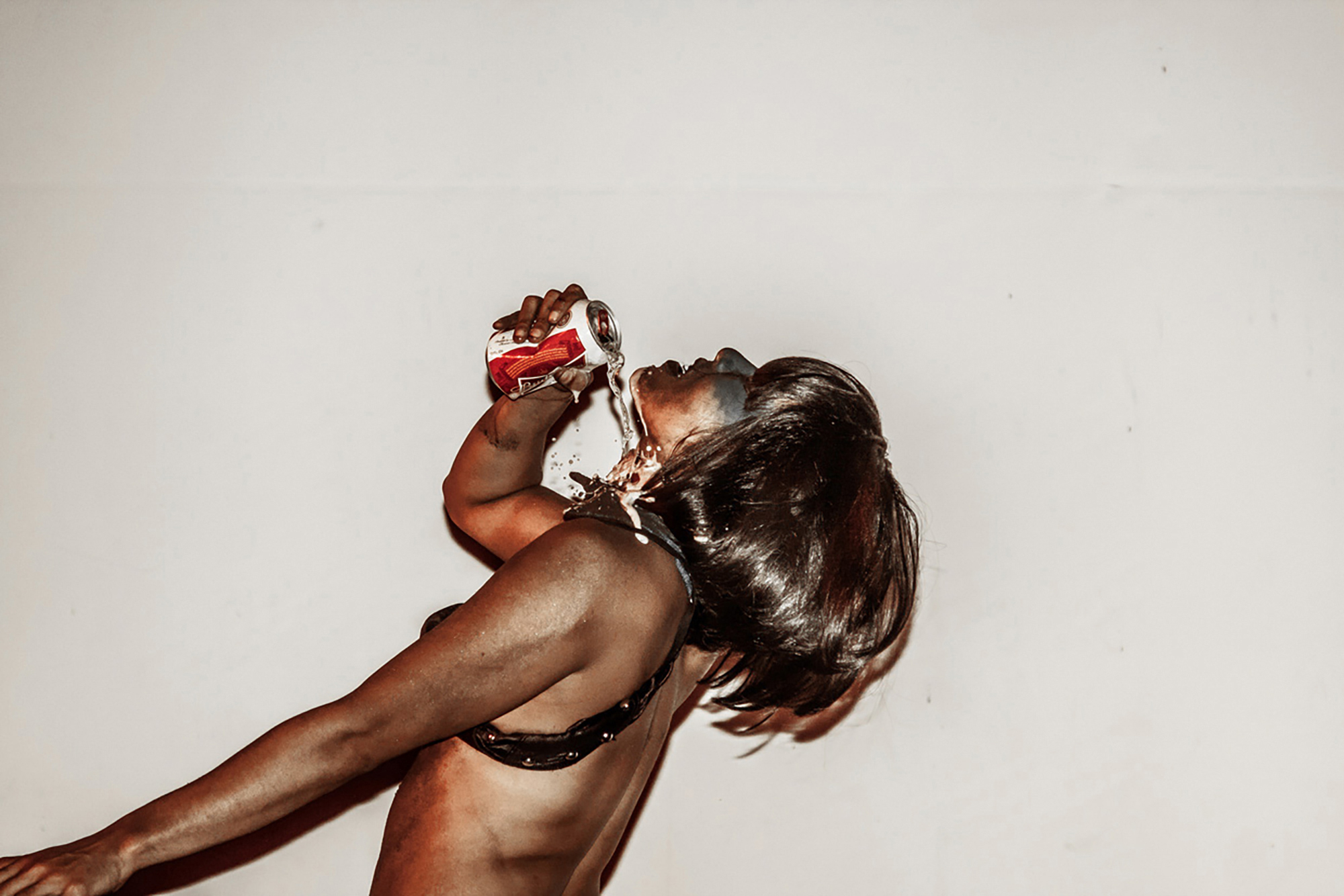 The profile of a brown woman in a black bra with short brown hair tilts her head upward. She hold a beer can with her hand and pours the beer onto her neck.