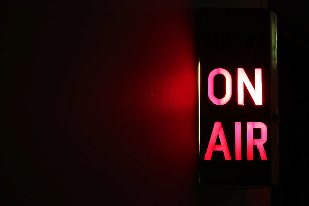 On the right side of the image an “On Air” sign is lit up with red letters in a dark room. The wall next to the sign is slightly red from the glow of the letters.