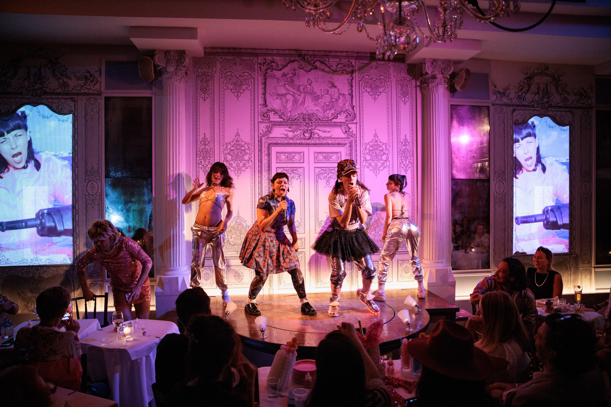 Liz and Bow are passionately singing into their microphones on stage with their dancers as a clip of Liz from Dame Leche is projected on both sides of the pink lit stage.