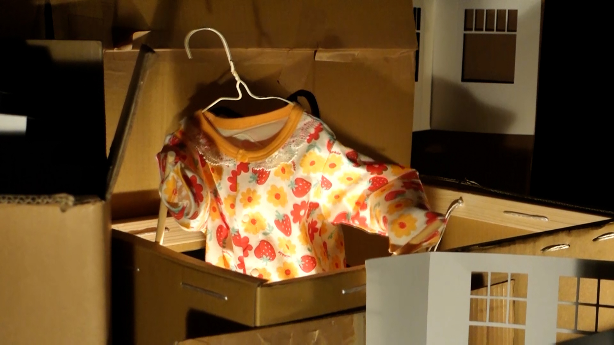 A puppet made out of a colorful baby clothes and a white metal hanger surrounded by cardboard boxes and paper cut-outs of windows.