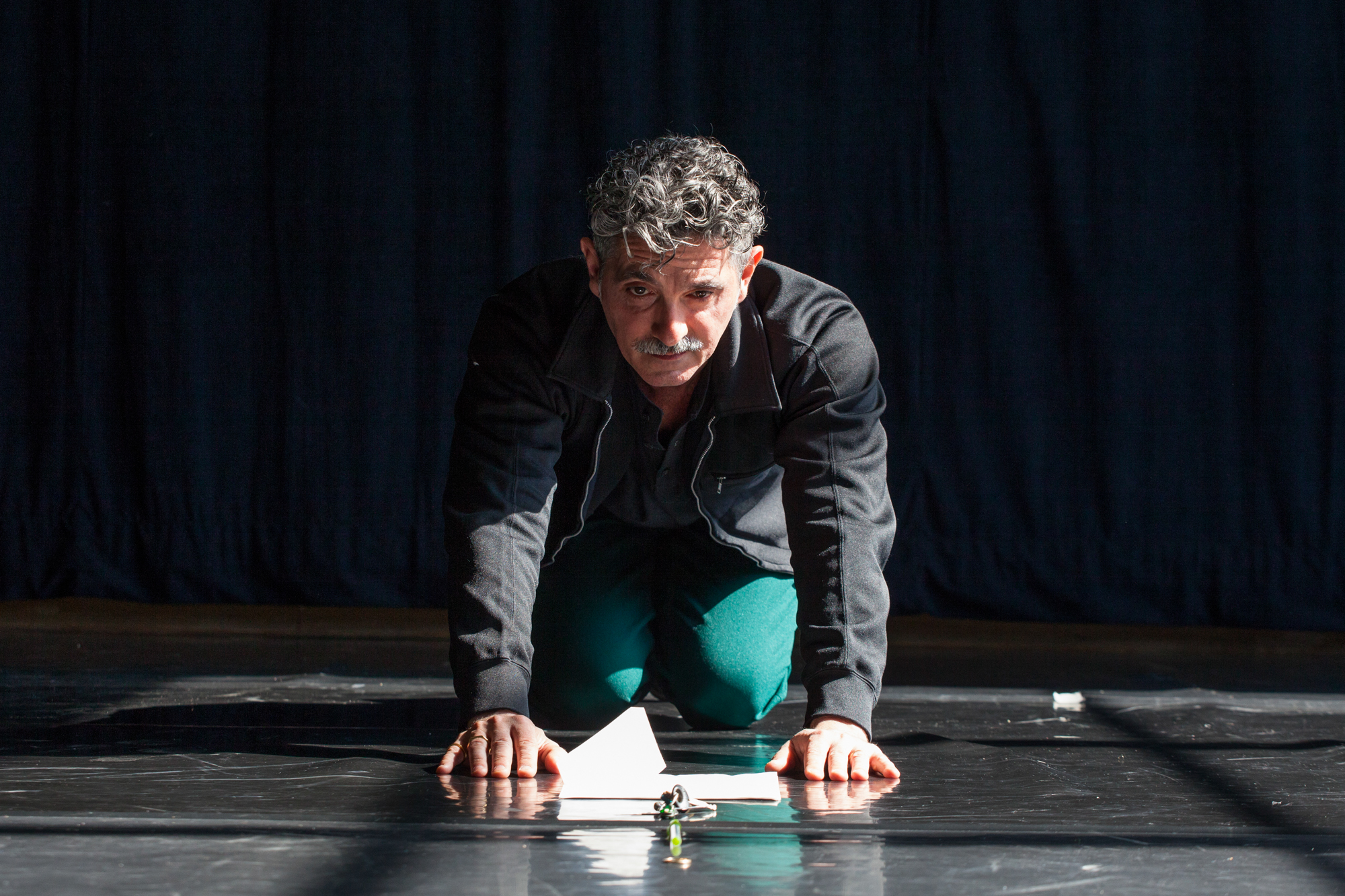 Shows a man, with salt and pepper hair, on his hands and knees contemplating a collection of seemingly ordinary objects on the floor.