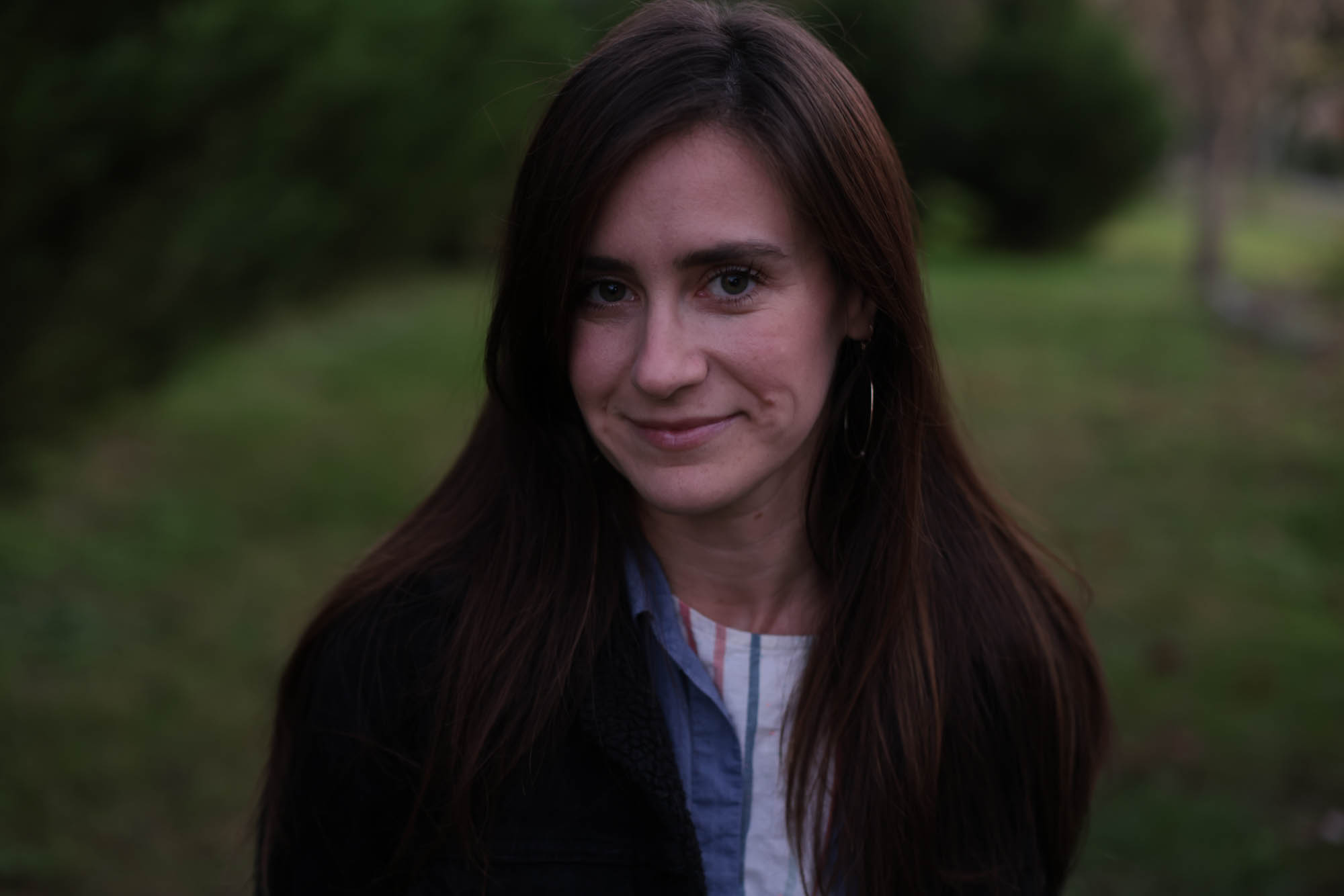 A caucasian woman with long brown hair, wearing a blue and white shirt and black jacket. Behind her is a rural landscape of trees and grass.