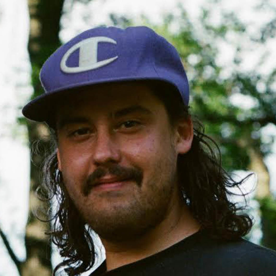 Anishinabe man with shoulder length black hair with a purple baseball cap on, smiling in the woods.