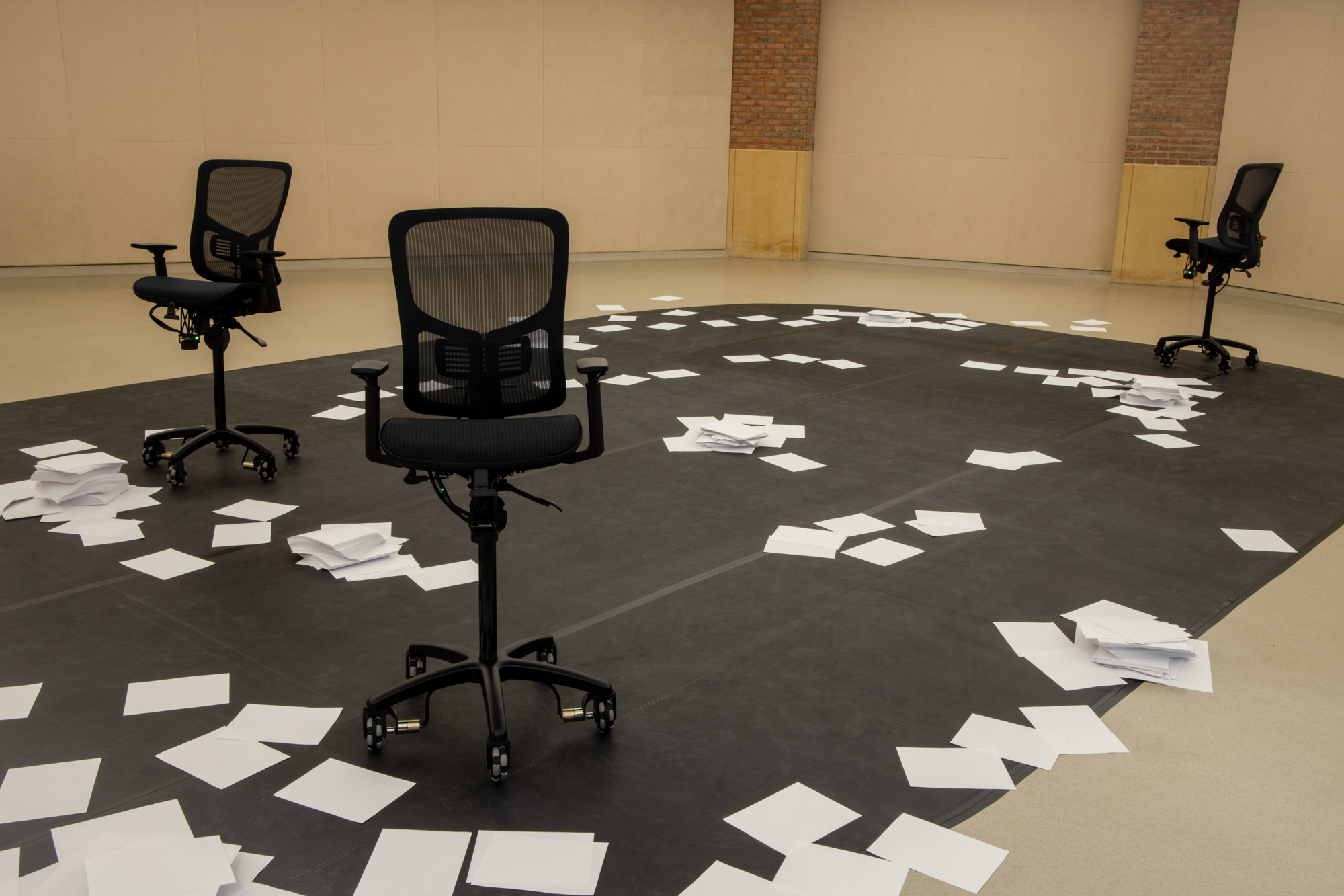 In a gallery, three black office chairs, modified with motorized wheels and electronic sensors, stand amidst a mess of white printer paper scattered in sheets and stacks on a black floor mat.