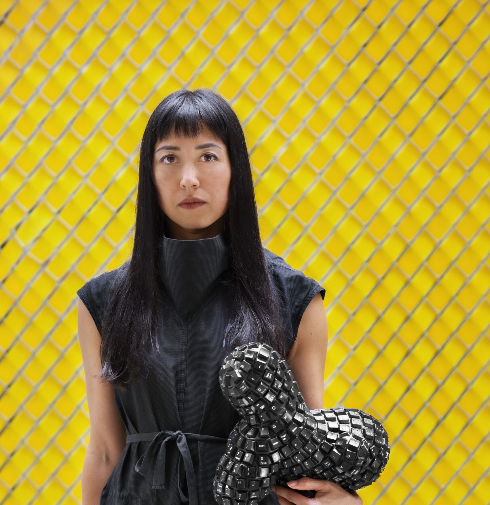 A woman in black stands in front of a bright yellow fence holding a sculpture covered in black QWERTY keyboard keys under her arm like a pet. She is of mixed-race Asian descent with long dark hair and light complexion and looks straight at the camera.