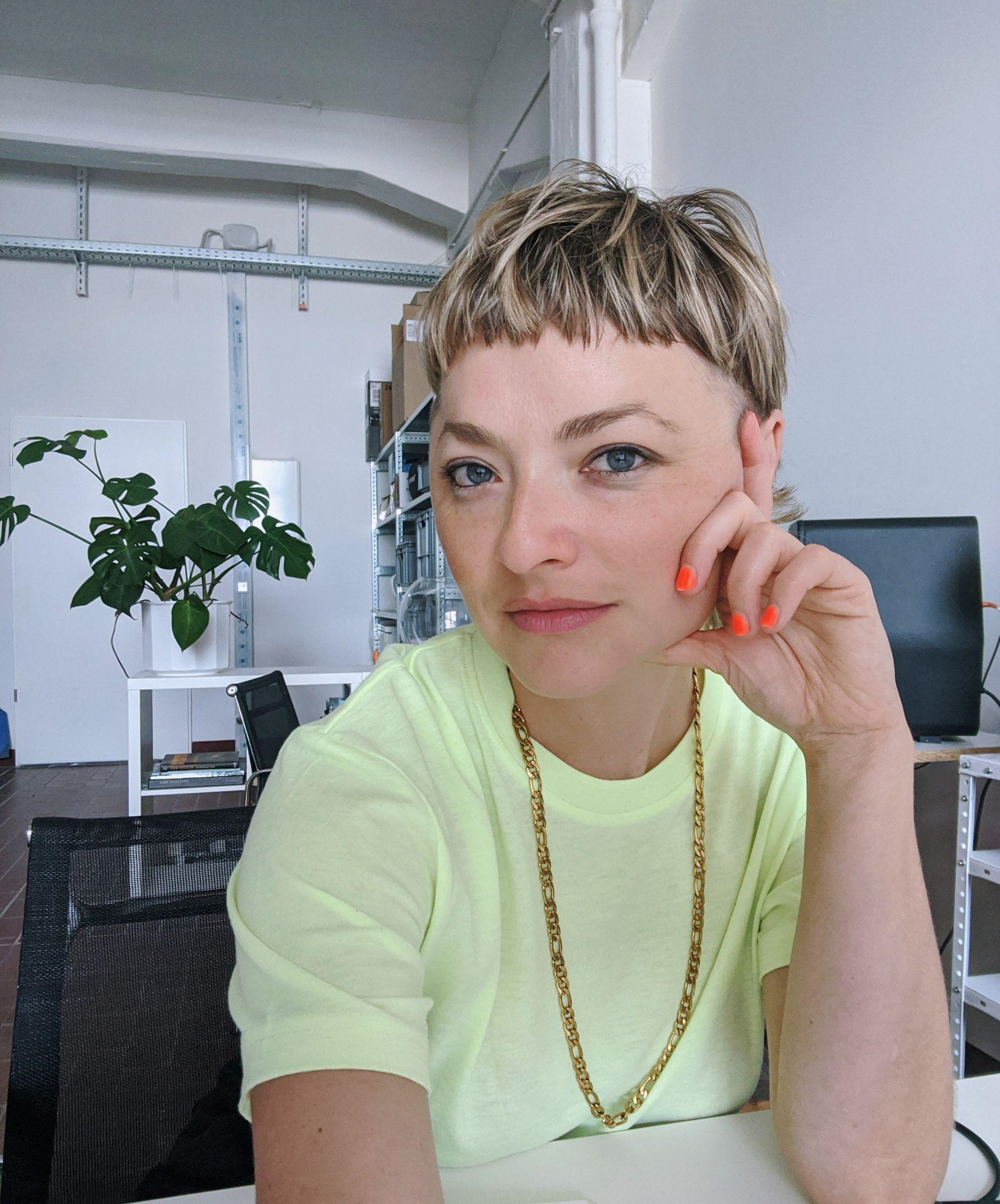 Caitlin looks at the camera in her studio. She has short blonde hair, blue eyes and neon orange fingernails, wearing fluorescent yellow t-shirt and a big gold chain.