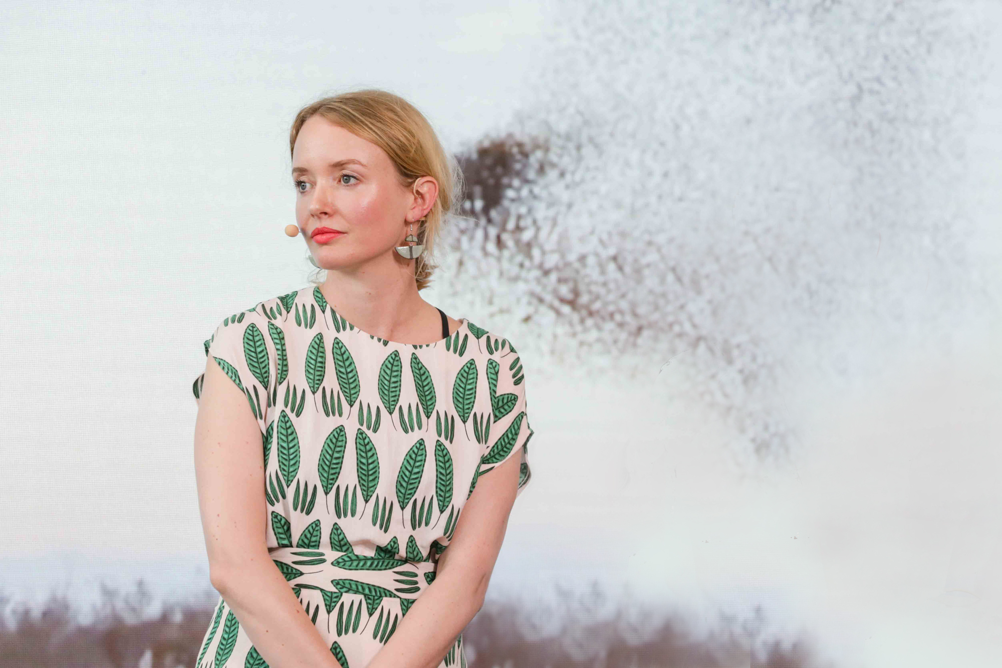 A blonde woman, in a green and white dress giving a talk and standing in front of a projection showing a flock of starlings.