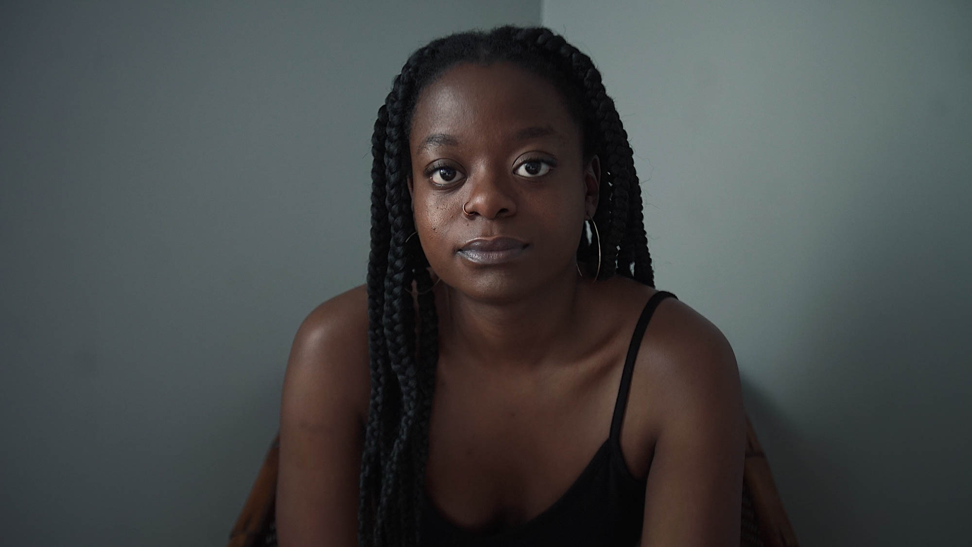 A woman with long black braids sits, leaning towards the camera. She's wearing gold hoops, a gold nose ring and a black camisole. She is sitting in a room with blue grey walls.