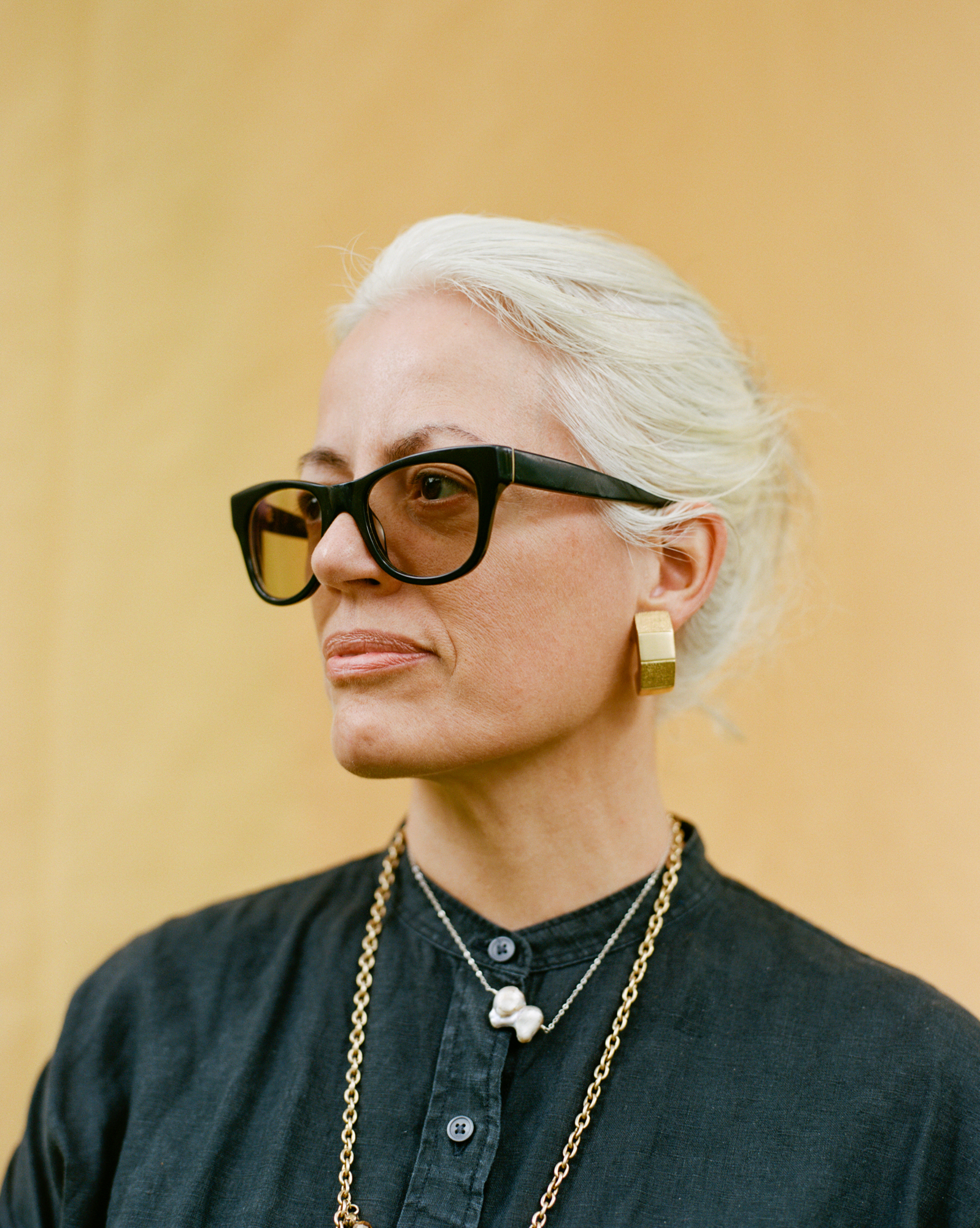 Photo of Yanira Castro, cropped just below her shoulders in front of a bright yellow background. She is looking off to the right, wearing black thick rimmed glasses, her white hair pulled back, a black linen shirt, a long golden chain, and her grandmother's gold cubist earrings.