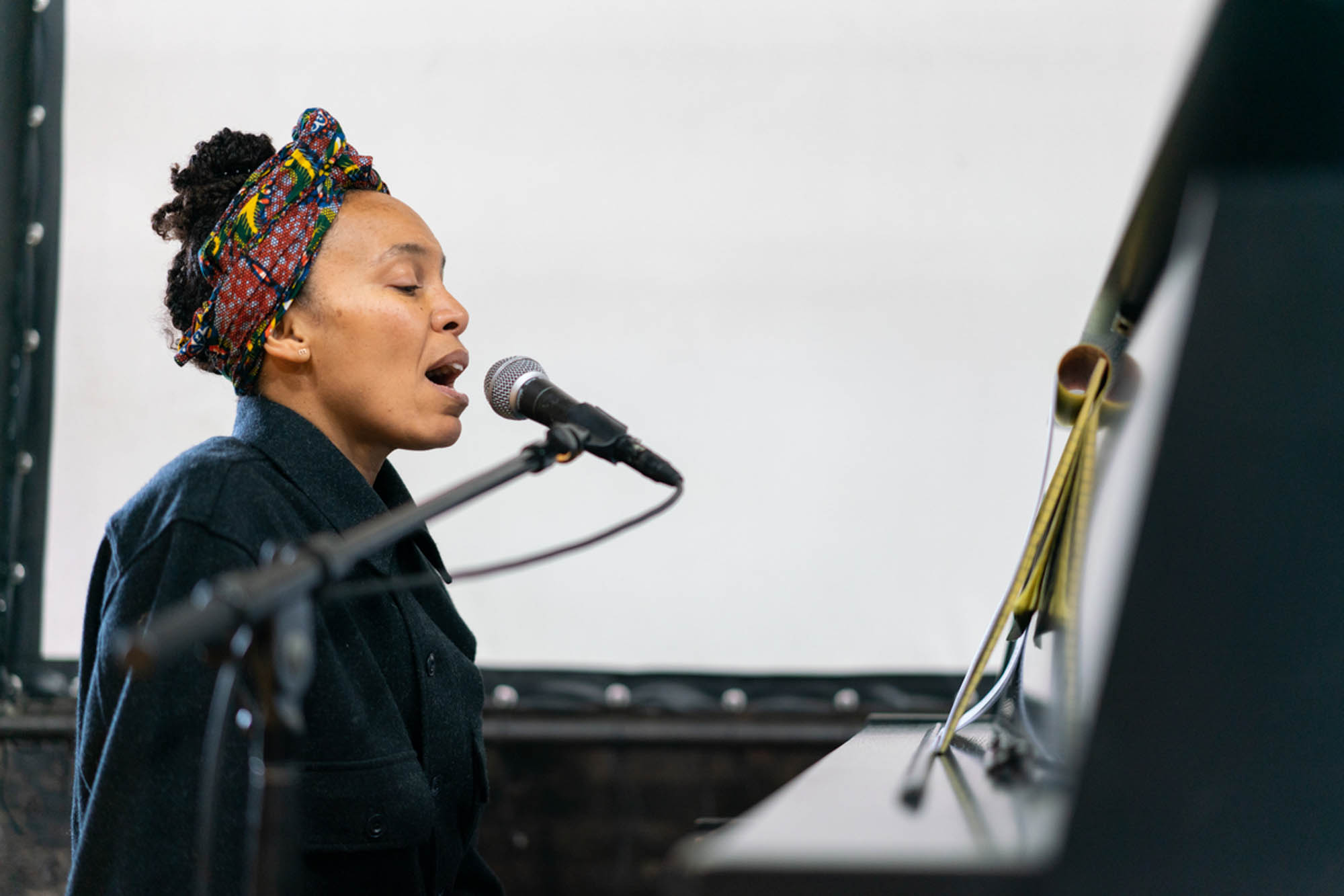 a black woman with African fabric wrapped on her head sings into a microphone at a piano gazing at her thoughts and intentions written on a yellow legal pad