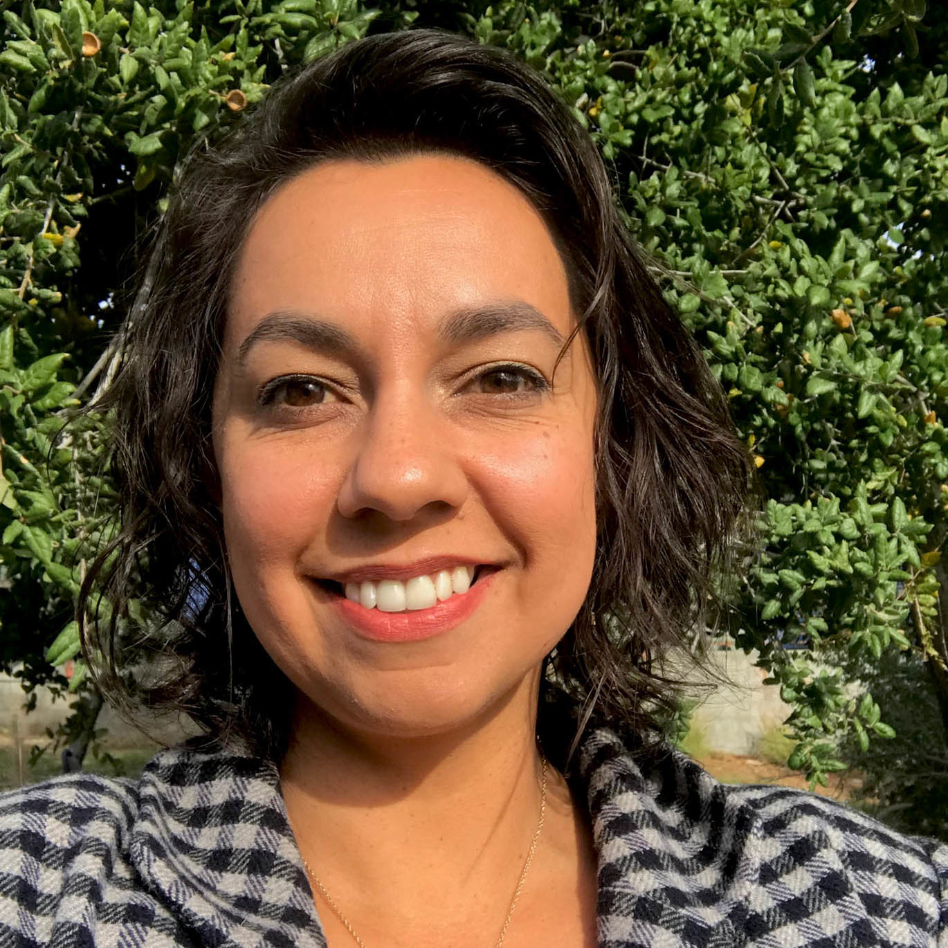 a woman in a checkered coat with wavy hair brown hair smiles toward the viewer in front of a California oak tree