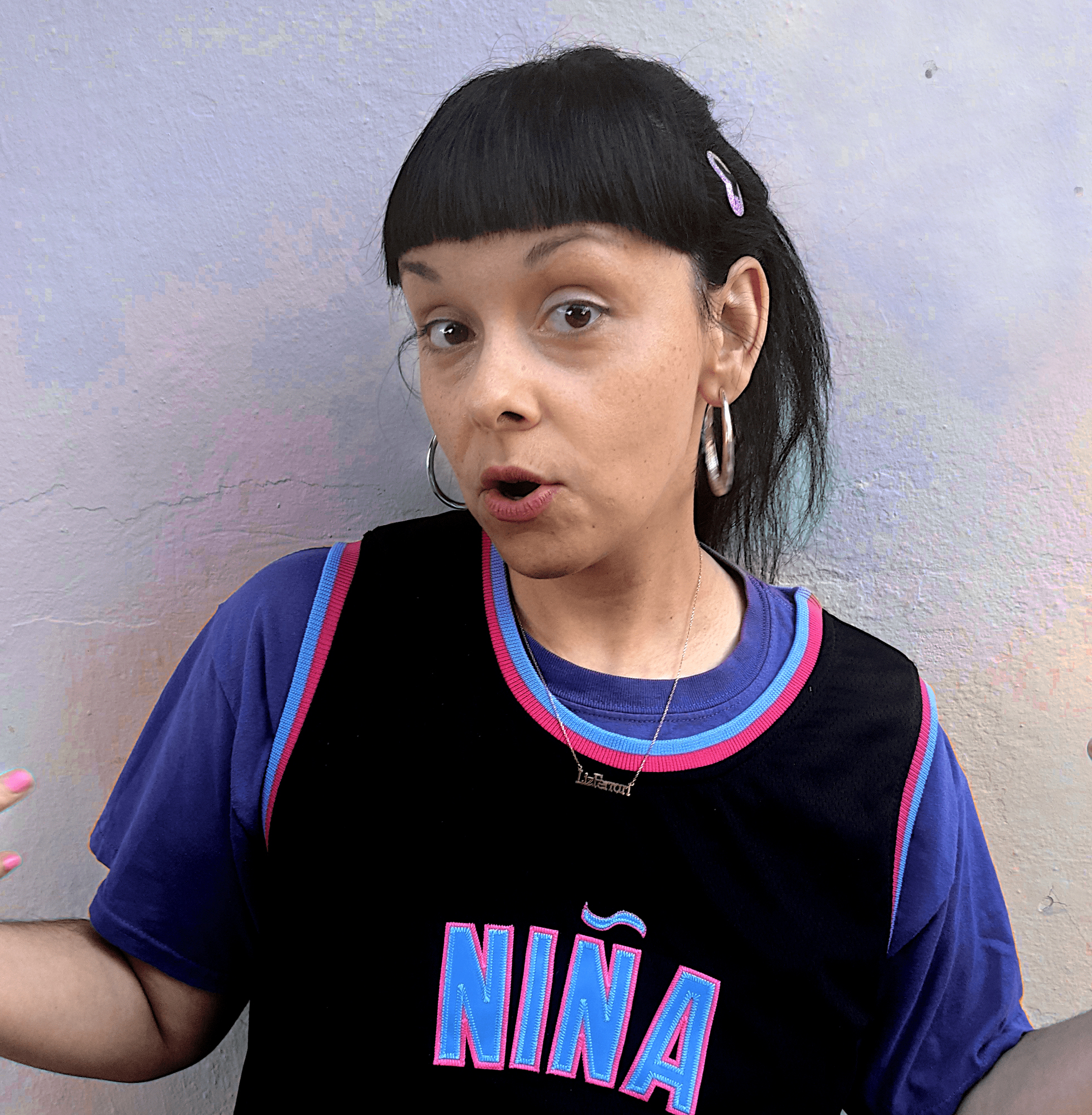 Image of artist Liz Ferrer mid sentence with a sassy affect, wearing silver hoops, a glitter heart clip, a small gold necklace that says