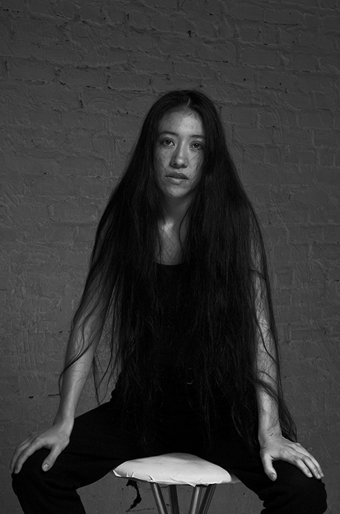A black and white image of a woman with long hair with a deep frontal gaze is seated on a high stool.