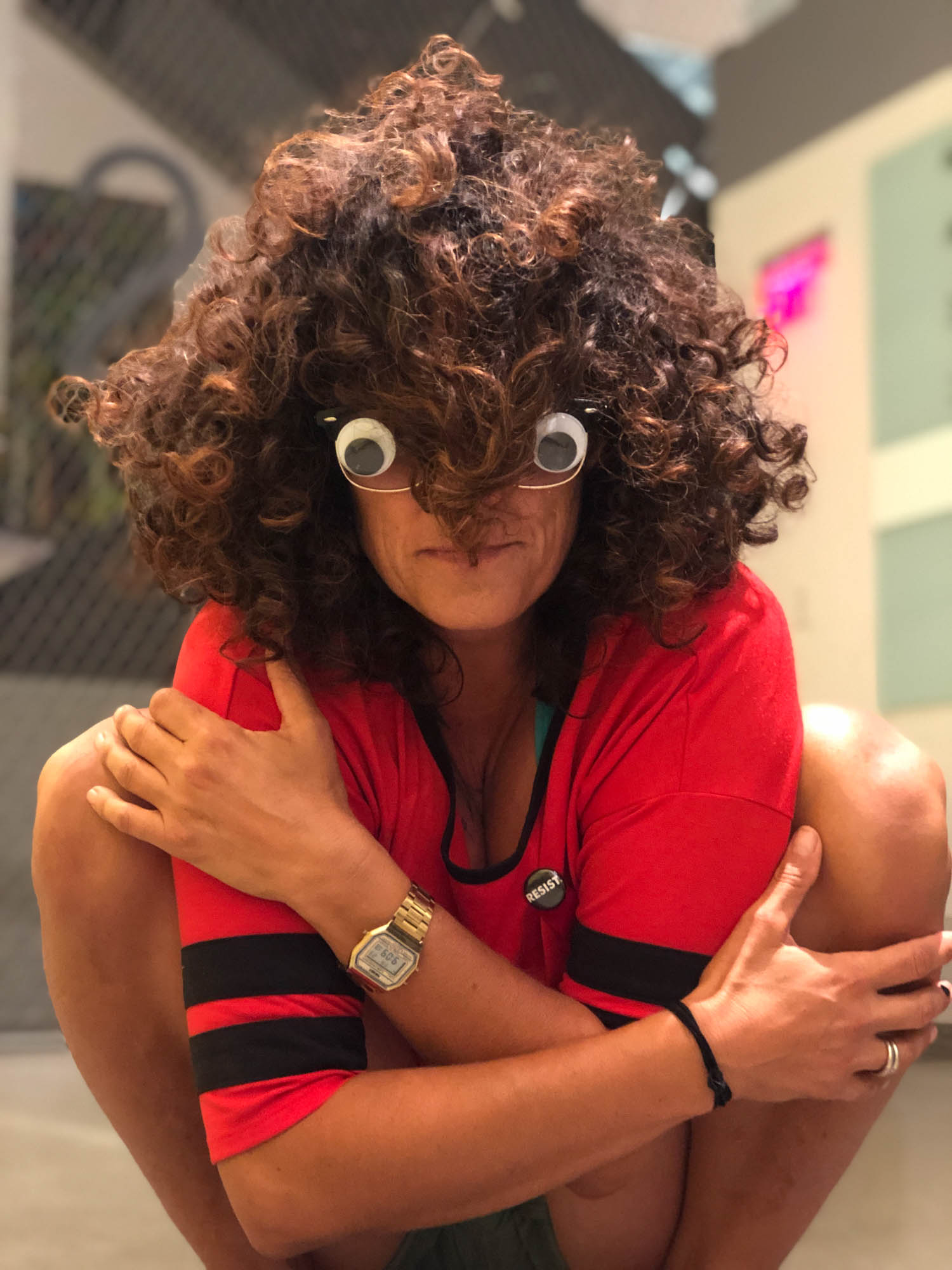 Woman with wild hair, arms crossed, and googly eyes stuck to her glasses in a deep squat, emanating light and exuberance