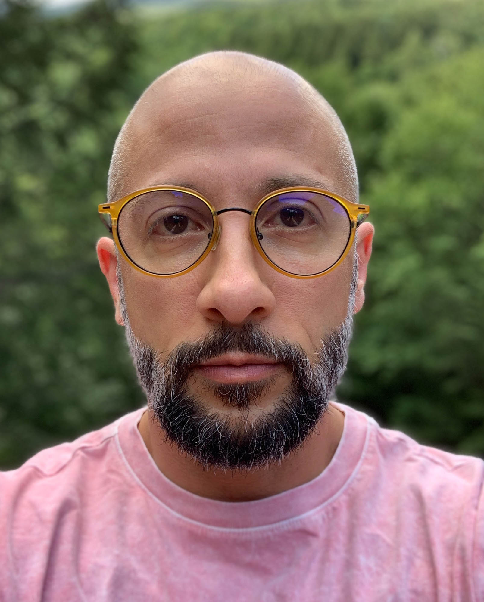 An Ashkenazi Jewish trans-masculine person is looking into the camera. He has a salt and pepper beard, yellow rimmed glass, and wears a mottled pink shirt. In the background is a lush out-of-focus forest.