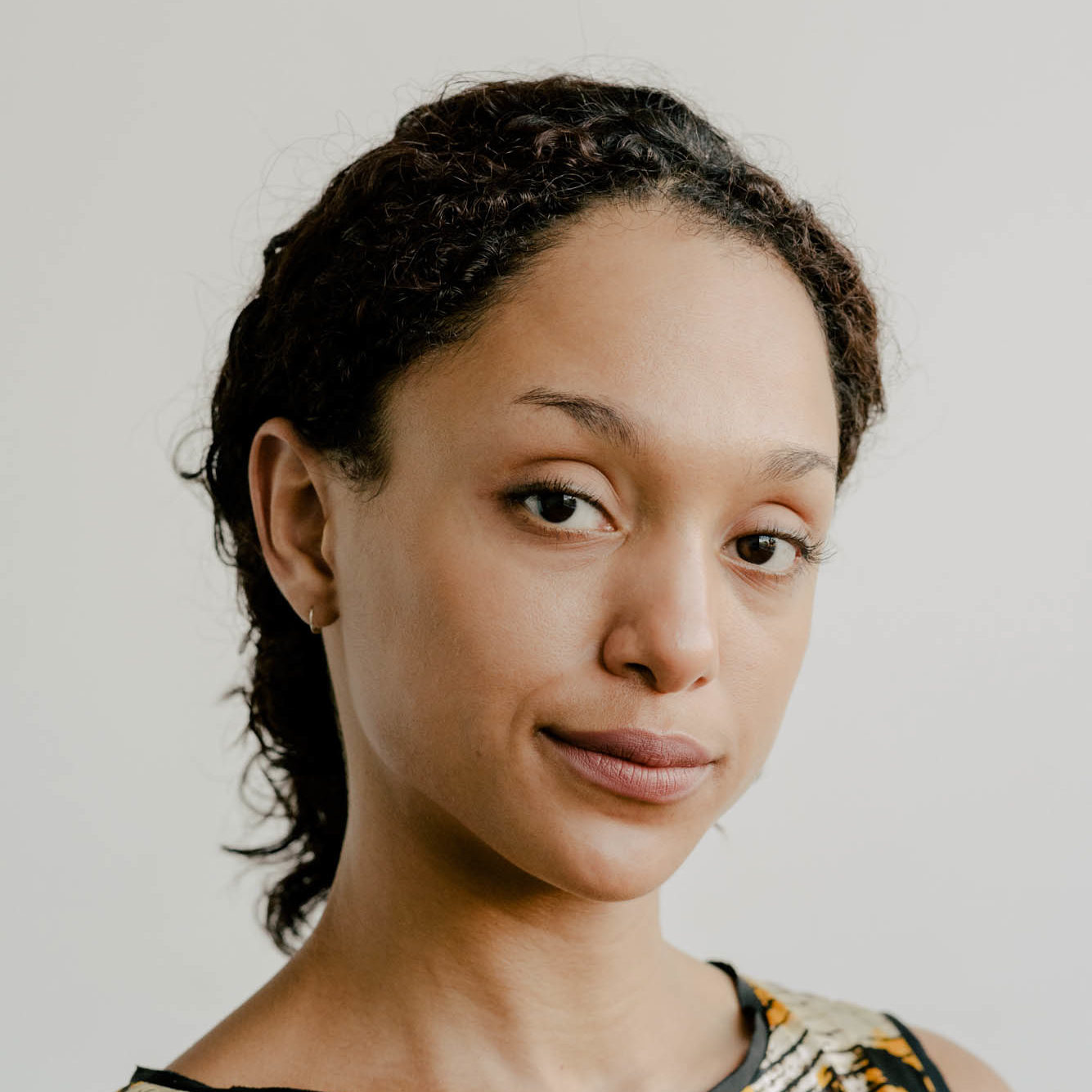 Black queer femme with hair tied back at the nape of her neck wearing a yellow and white floral dress with black trim looks sideways at the camera with chin tilted up.