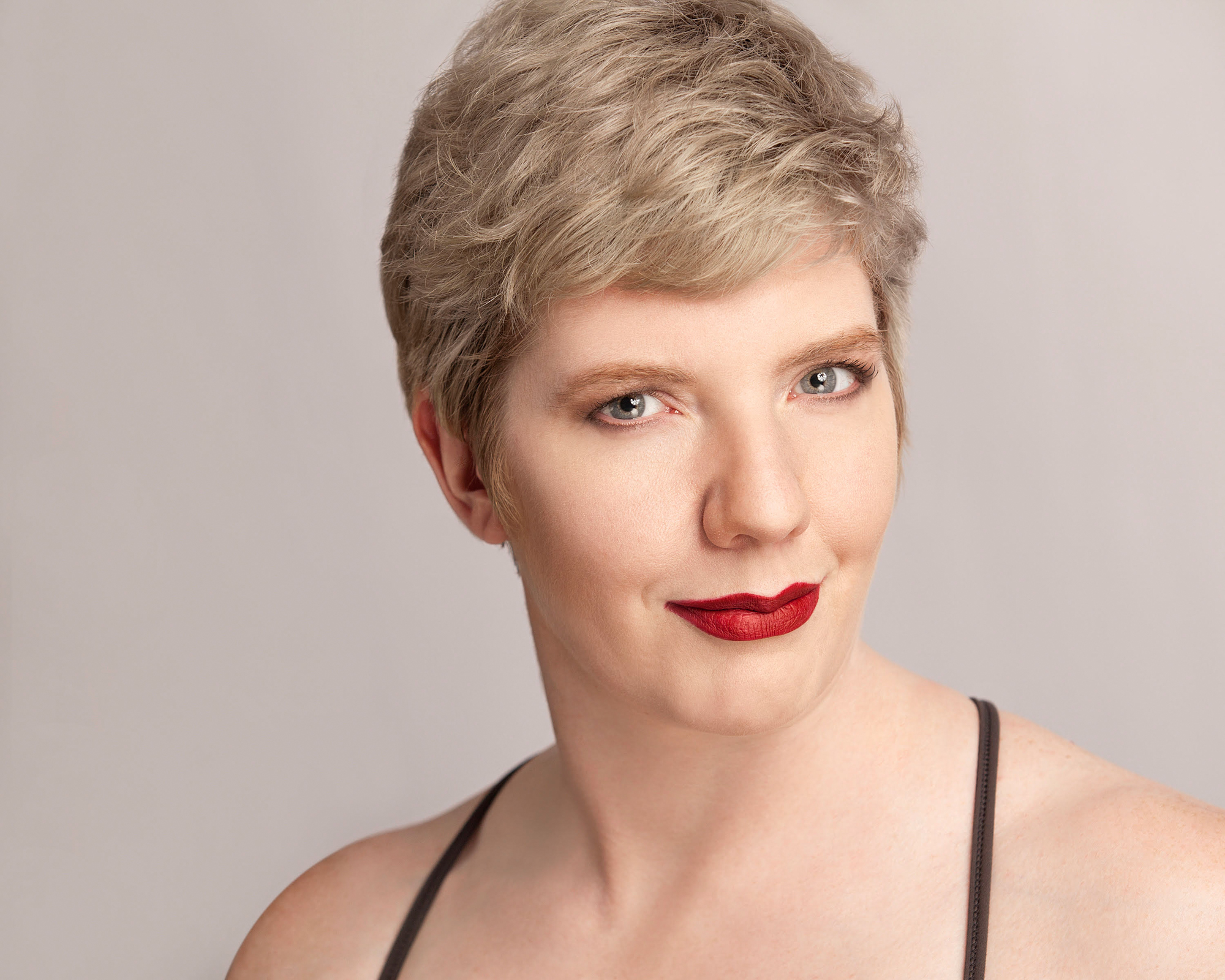 Laurel, a woman with pale skin and eyes and pixie-cut silvery hair, looks directly at you with an impish gaze and slight smile. She wears a grey leotard and bright red lipstick.