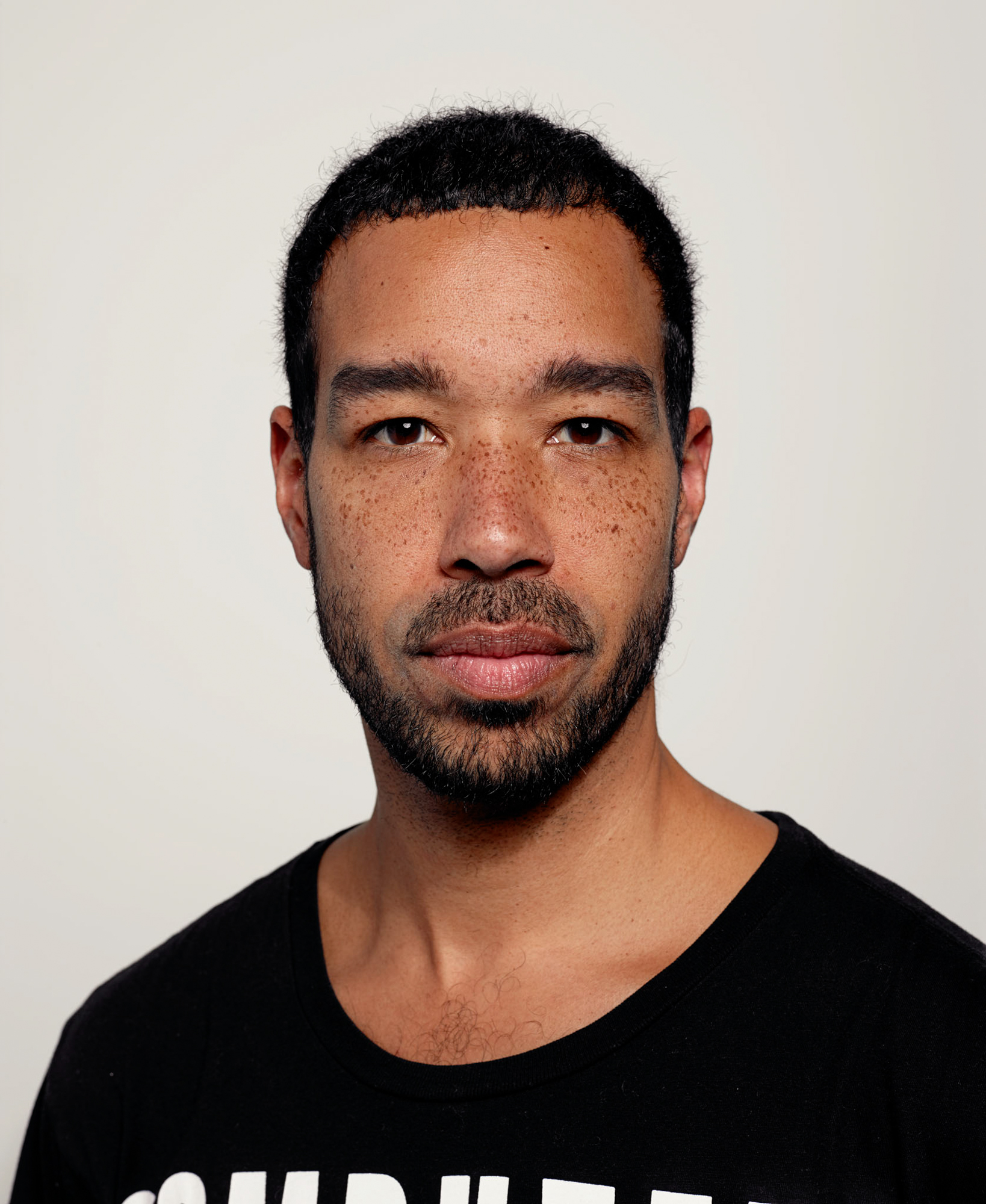 Rashaun is a light-skinned Black man with freckles, dark, almond-shaped eyes, dark, close-cut hair, and some facial stubble, wearing a black t-shirt with white writing.